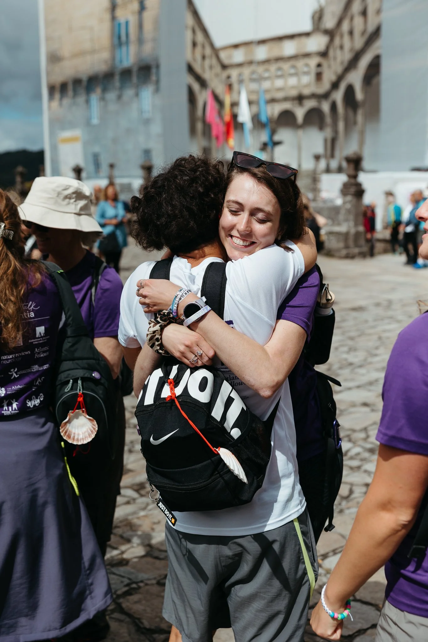 Two women hugging each other in a public square, surrounded by other people, with historic buildings and flags in the background.