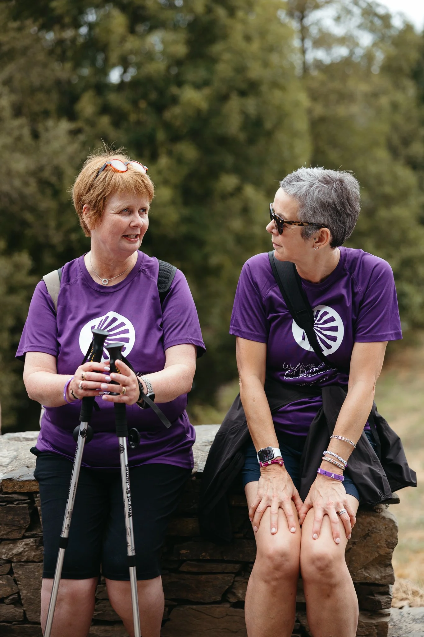 Two women in purple t-shirts sitting on a stone wall outdoors, engaged in conversation against a background of trees.