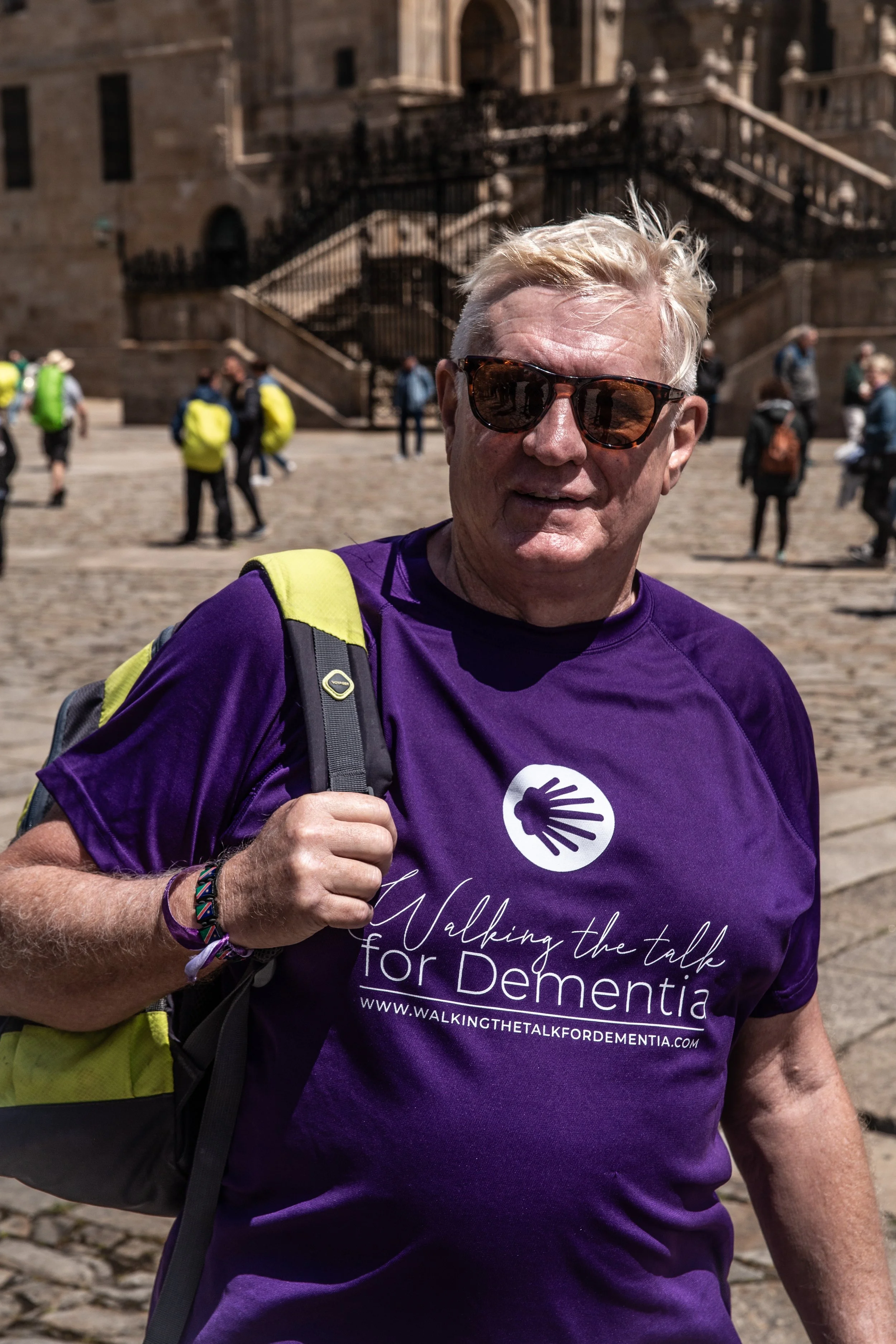 A man with blond hair wearing sunglasses, a purple T-shirt with a logo and the text "Walking the talk for Dementia," and a backpack, standing outside on a cobblestone square with a historic building and several people in the background.