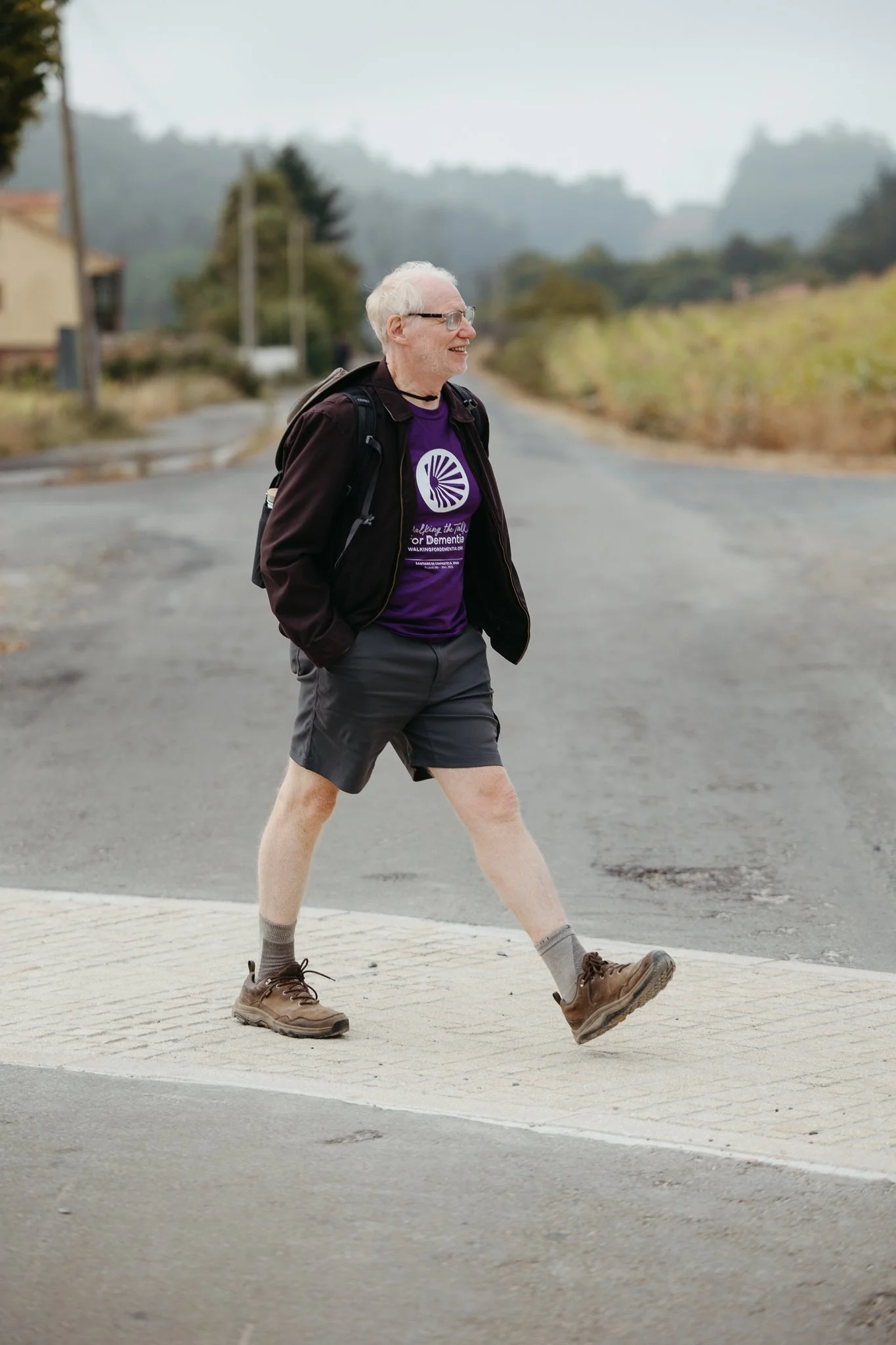 Older man with white hair, glasses, wearing a purple T-shirt, black jacket, gray shorts, hiking boots, and carrying a backpack, crossing a street.