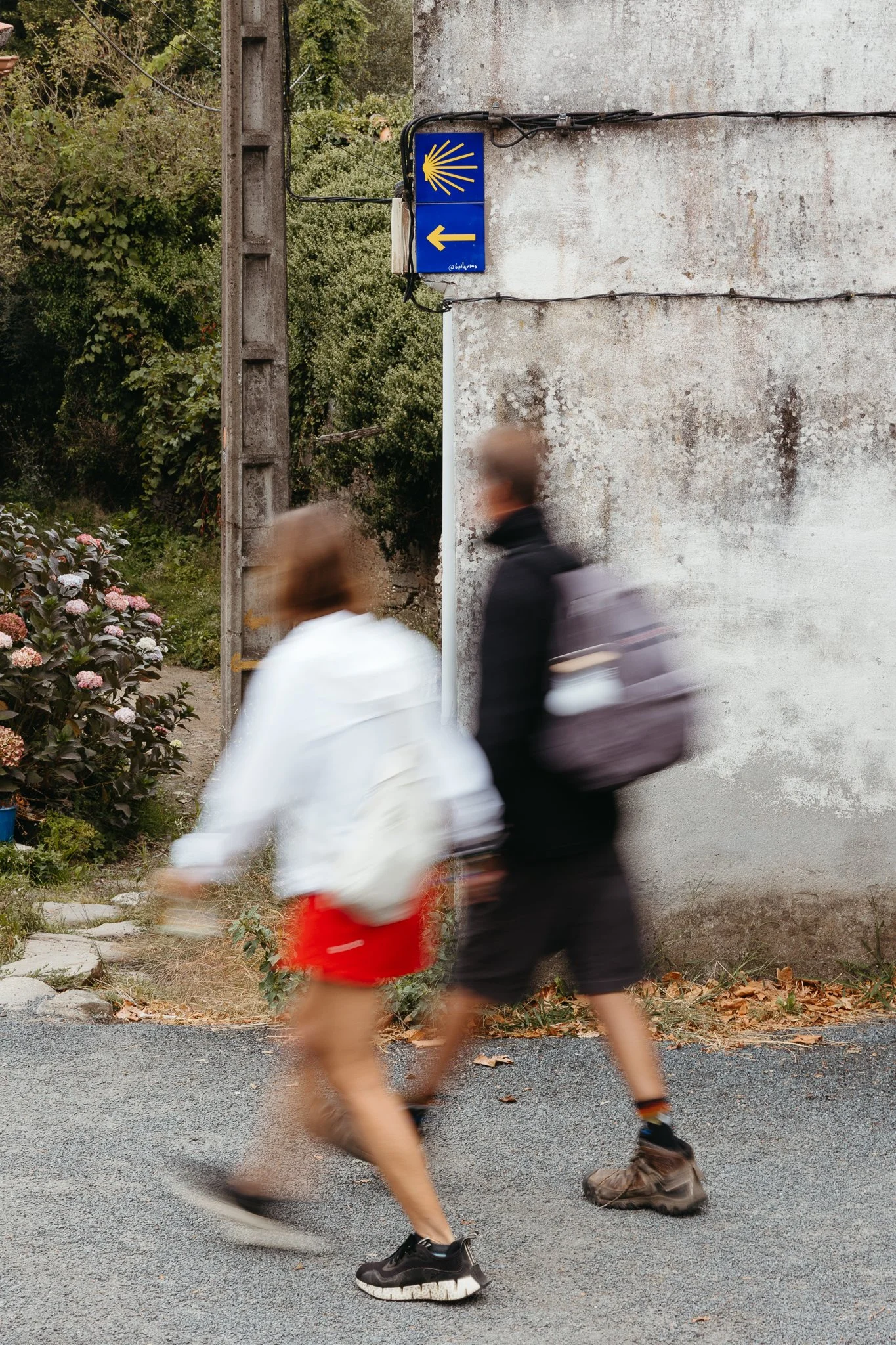 A blurry image of two people walking past a wall with a blue sign showing a yellow shell and an arrow pointing left, indicating a Camino de Santiago route.