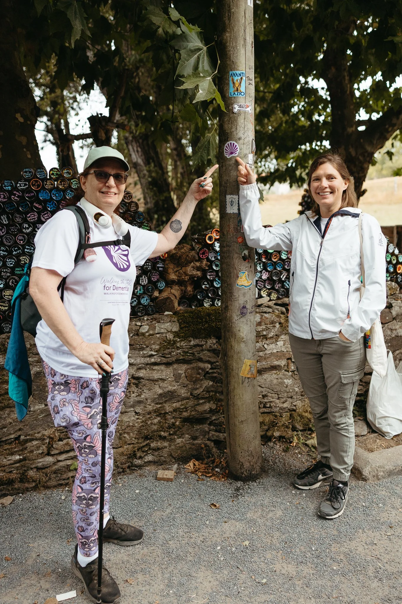Two women standing outdoors next to a wooden post with stickers, smiling and pointing at stickers. One woman is wearing sunglasses, a white cap, a white shirt, patterned pants, and carrying a walking stick. The other woman is in a light-colored jacke