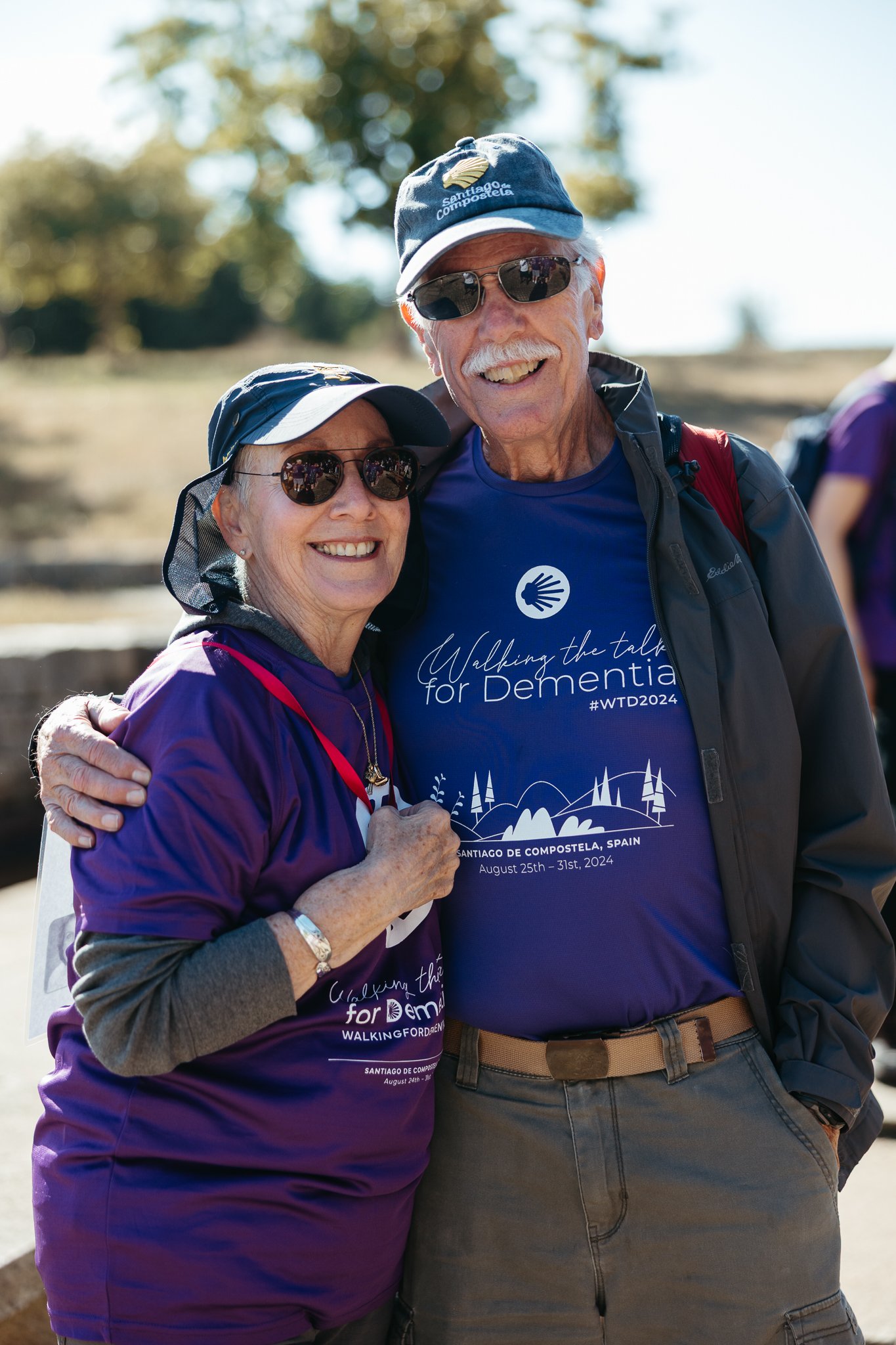 A smiling elderly woman and man outdoors wearing matching purple shirts for a walkathon event in Santiago de Compostela, Spain, with trees and blue sky in the background.