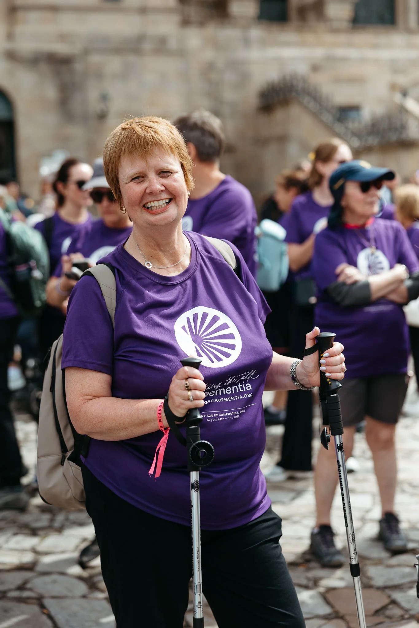 A smiling woman holding trekking poles at a walking event, surrounded by other participants in purple shirts.