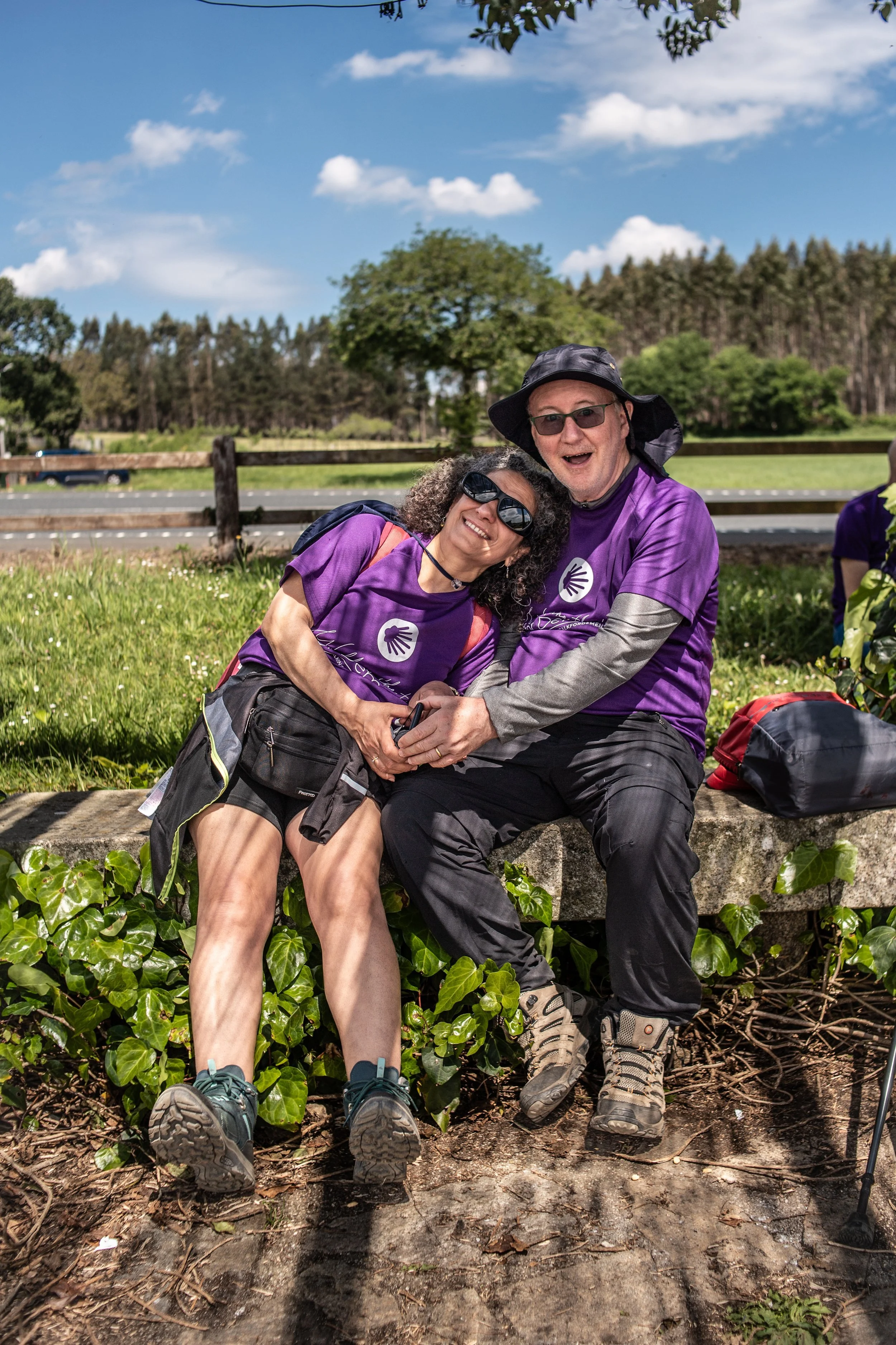 Two people sitting on a stone bench outdoors, smiling and holding hands, wearing purple shirts and sunglasses, surrounded by greenery on a sunny day.