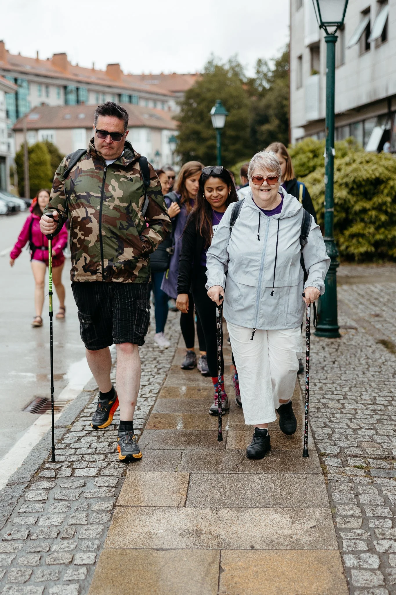 A group of people, including an elderly woman and a man, walking on a cobblestone sidewalk in a residential area with lampposts and buildings, some carrying walking sticks.