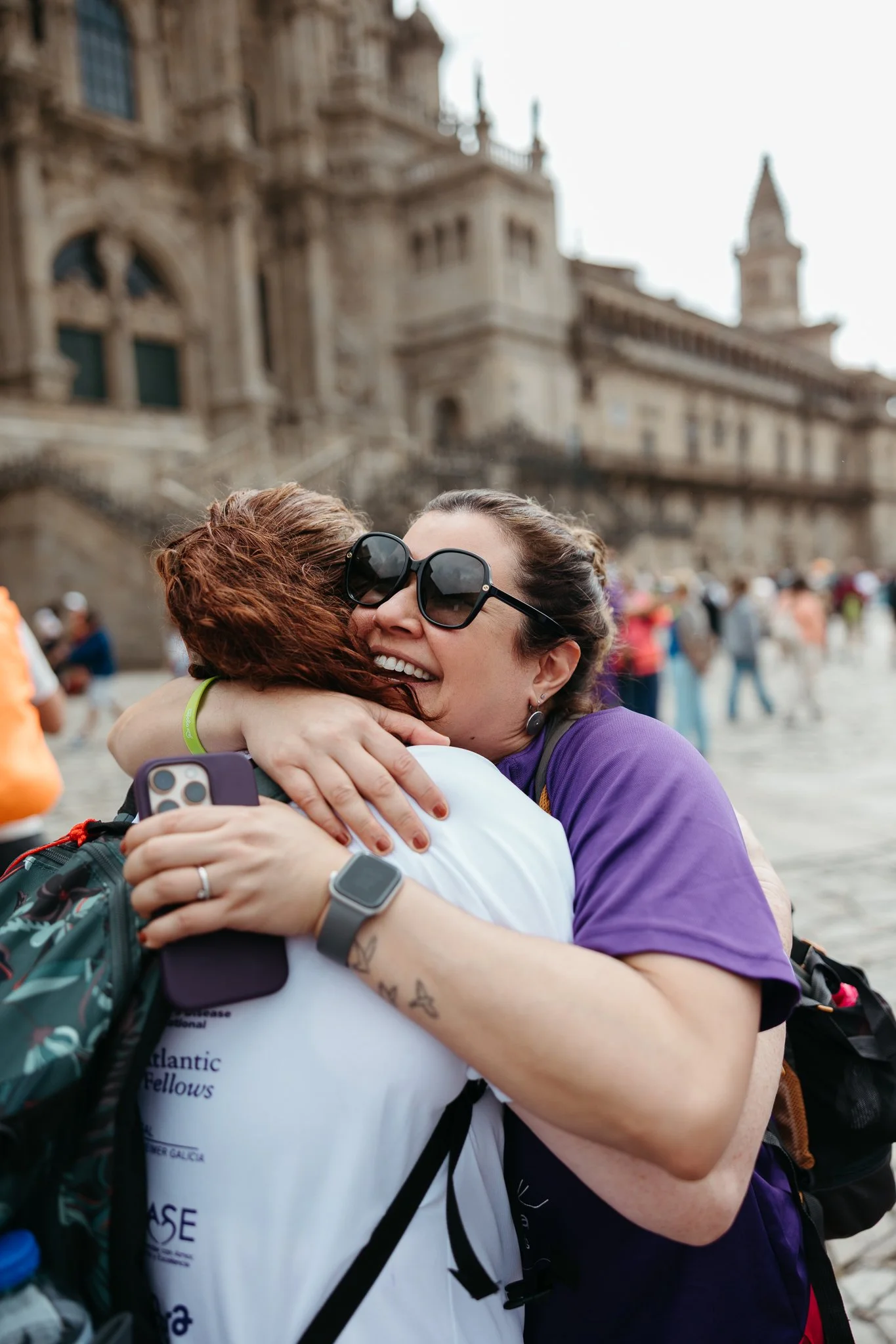 Two women hugging outdoors in front of a historic building, with one woman wearing large sunglasses and a purple shirt, smiling.