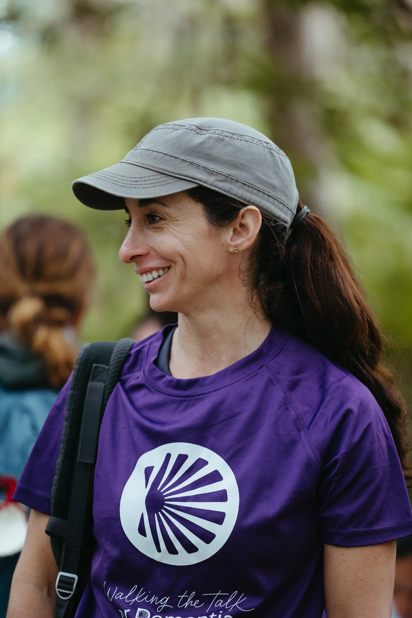 A smiling woman wearing a gray cap and a purple T-shirt with a logo, outdoors in a wooded area.