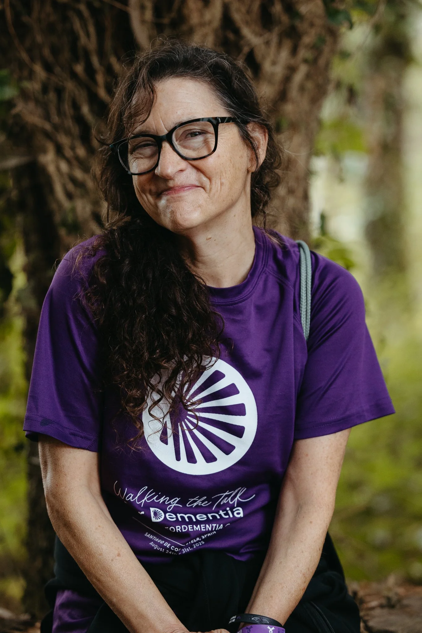 A woman with glasses and long curly hair, wearing a purple T-shirt with a white logo and text, sitting outdoors in front of a tree.