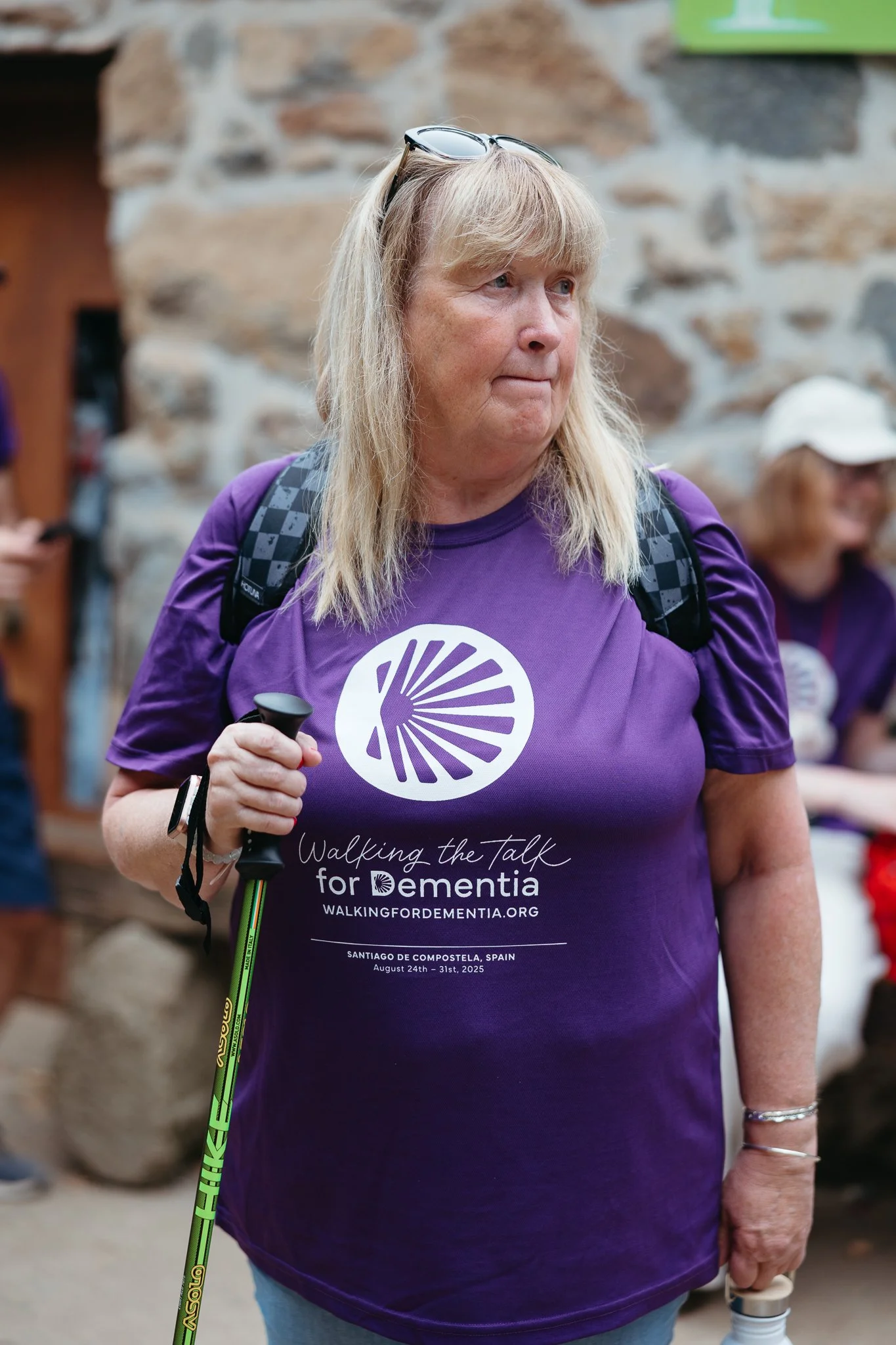 A woman wearing a purple t-shirt participating in a dementia awareness walk, holding a green walking stick, with glasses on her head, in an indoor setting with a stone wall background.