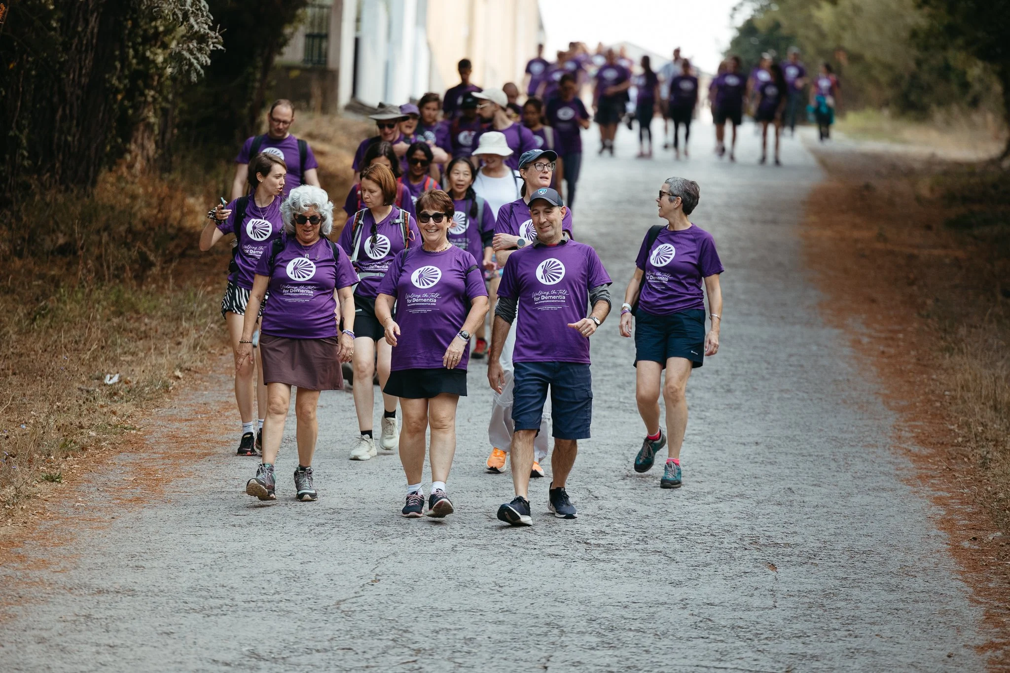 Group of people walking outdoors on a dirt path, wearing matching purple t-shirts supporting a cause for dementia awareness.