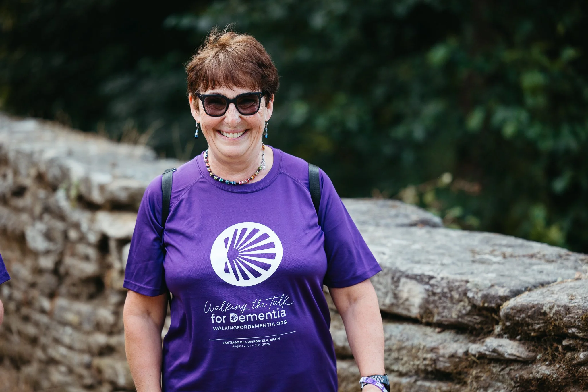 Smiling woman in sunglasses, wearing a purple t-shirt supporting a dementia awareness walk, standing outdoors with a stone wall and greenery in the background.