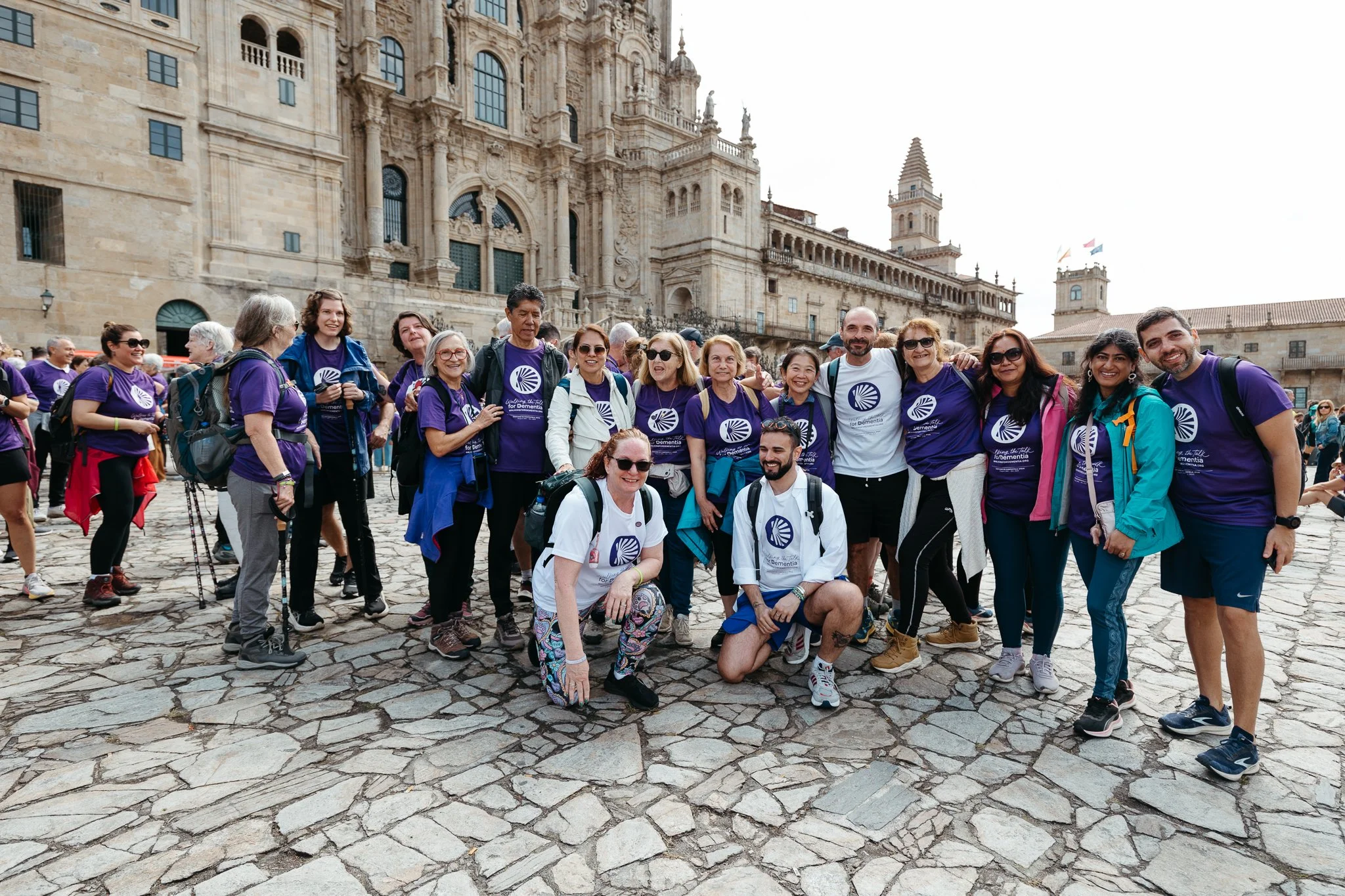 Group of people posing for a photo in front of historic building, wearing matching purple and white T-shirts with a logo, on cobblestone square.