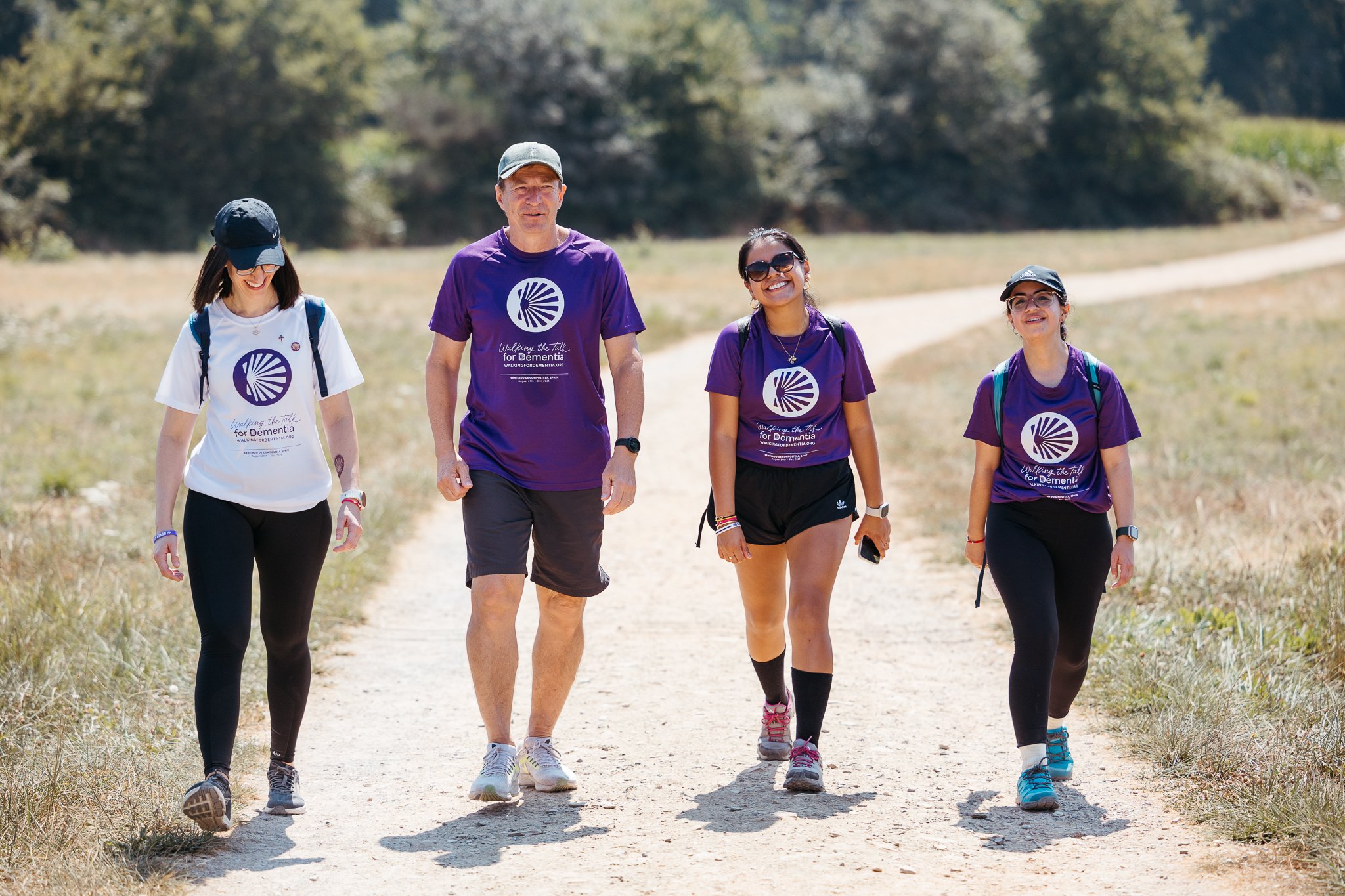 Four people walking outdoors on a sunny day, wearing purple shirts promoting a dementia awareness walk, with two women and two men.
