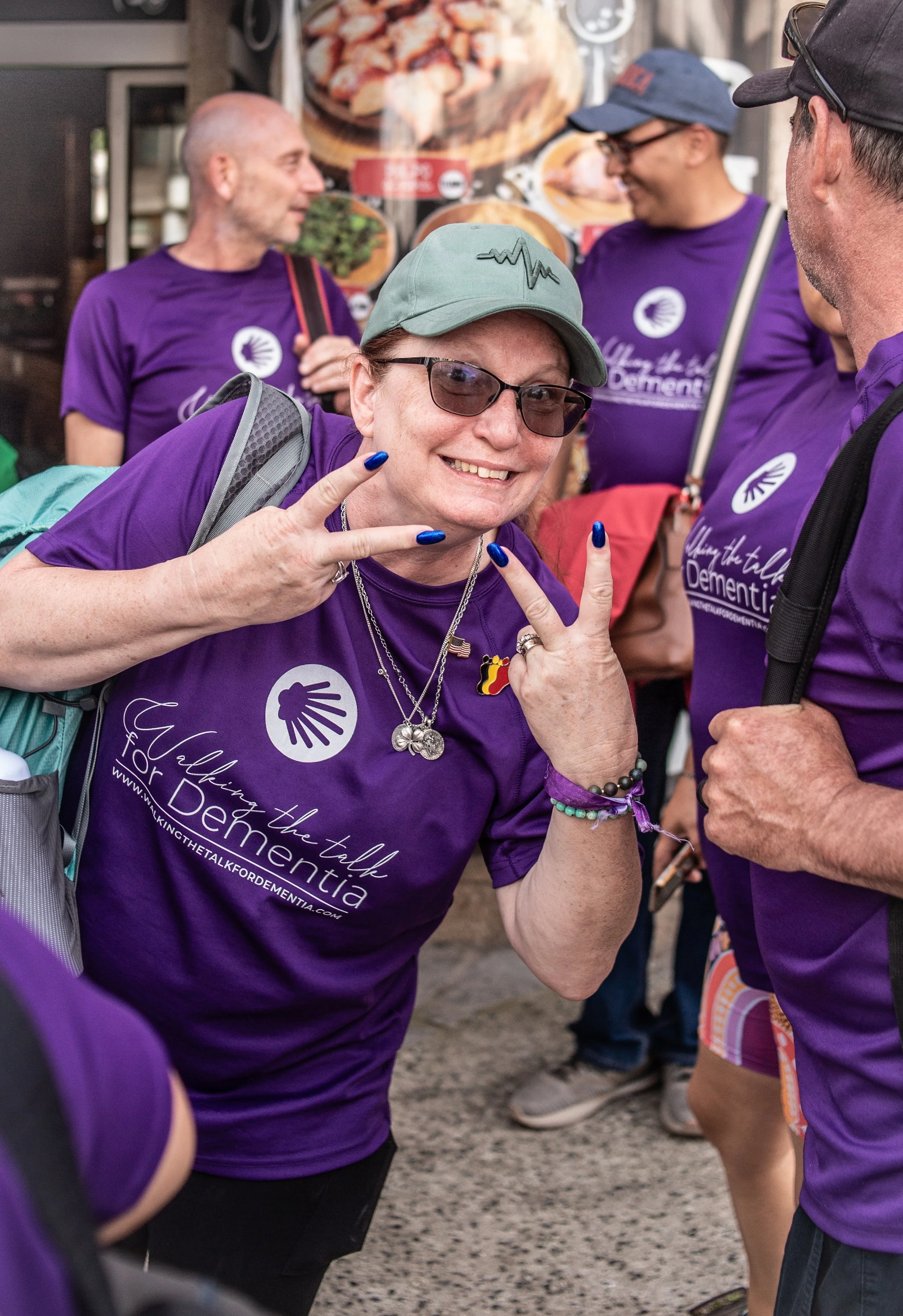 A woman wearing a purple T-shirt, sunglasses, a gray cap, and jewelry, smiling and making peace signs with both hands among a group of people in purple shirts during a charity event about dementia.