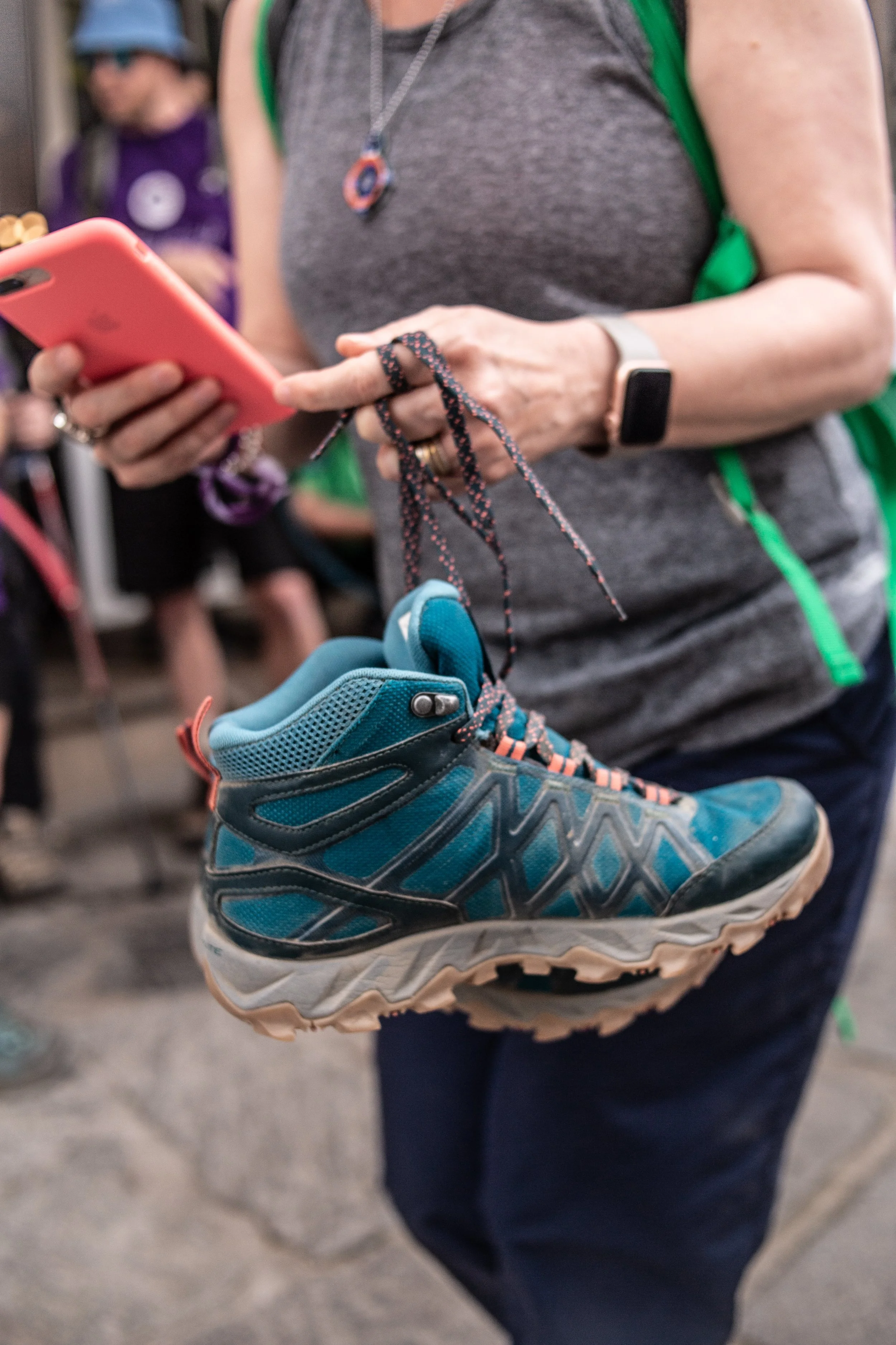 Person holding a turquoise hiking shoe and looking at a smartphone at a busy indoor or outdoor event.