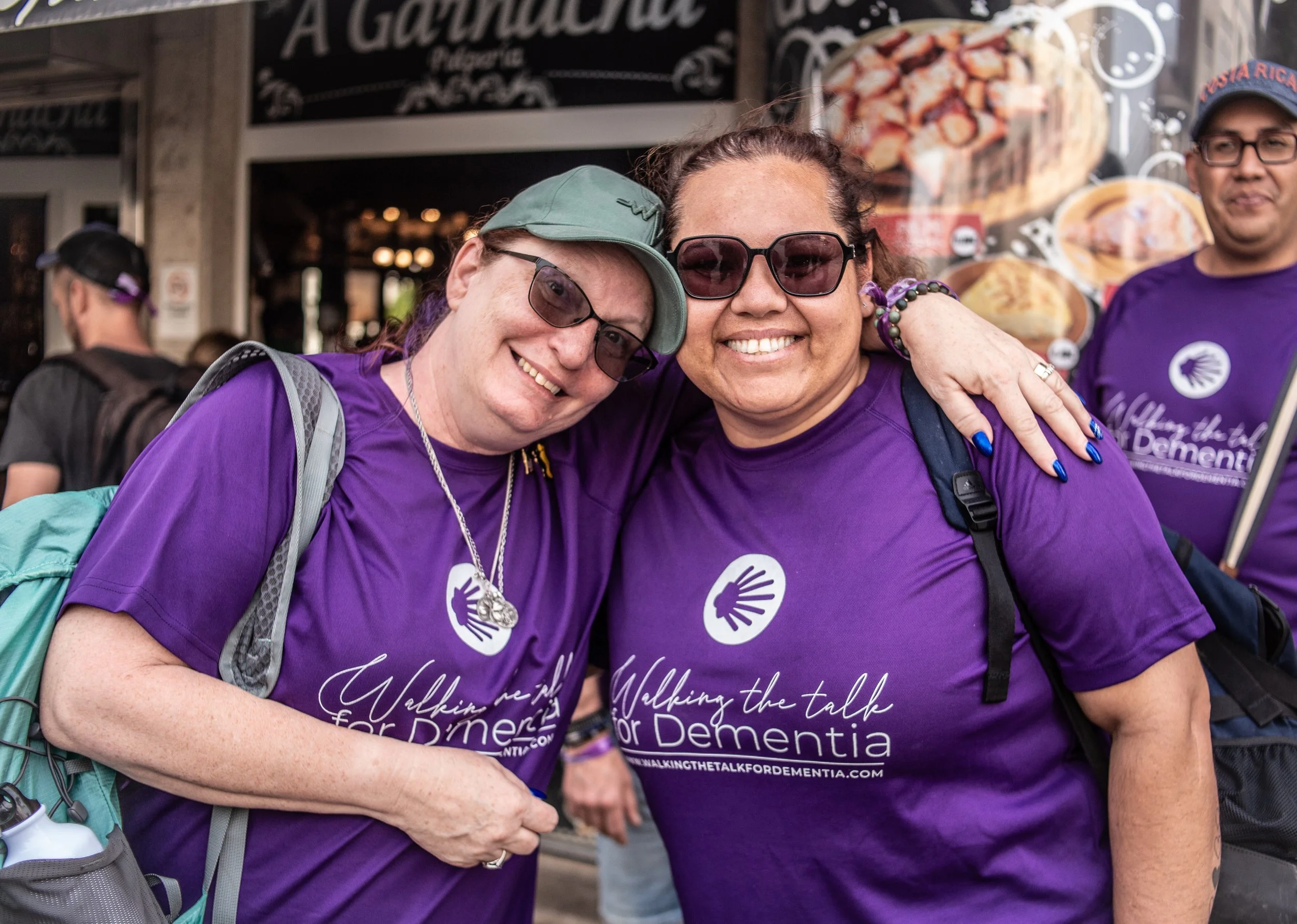 Two women wearing purple shirts with the logo and text 'Walking the talk for Dementia' are smiling and hugging in a crowd.
