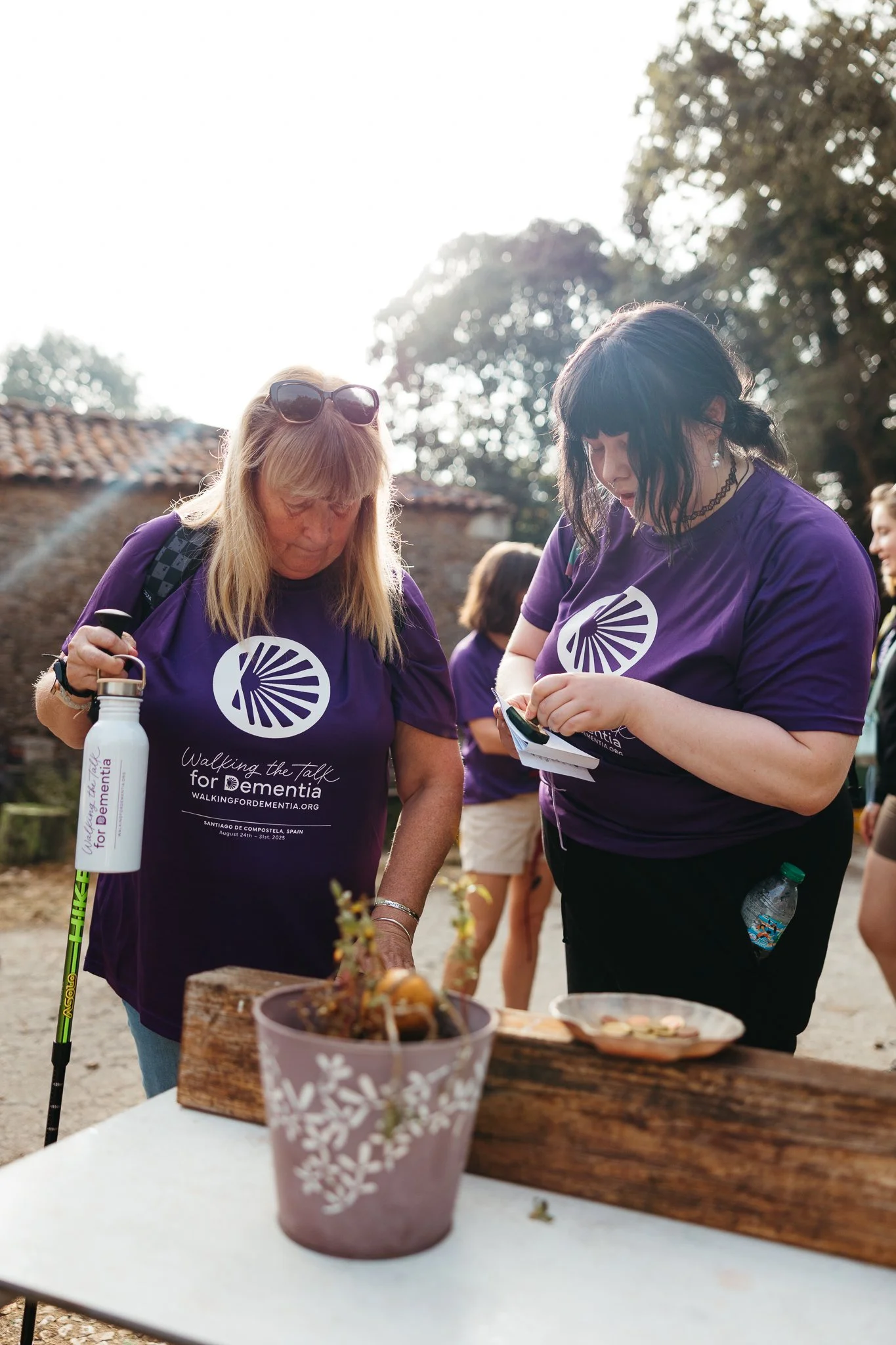 People participating in a dementia awareness walk wearing purple shirts, standing at an outdoor table with a potted plant, taking notes, and holding water bottles.