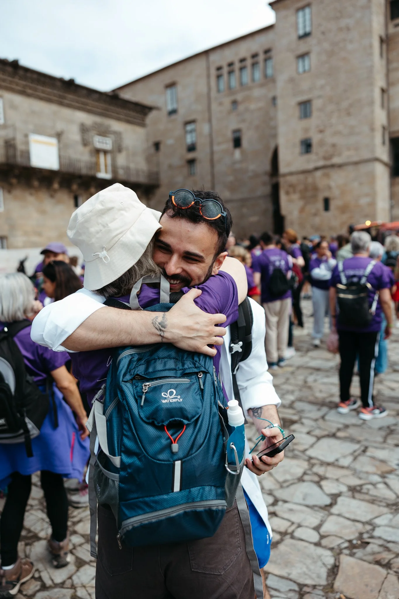 A man and woman hugging among a crowd of people outside a historic stone building.