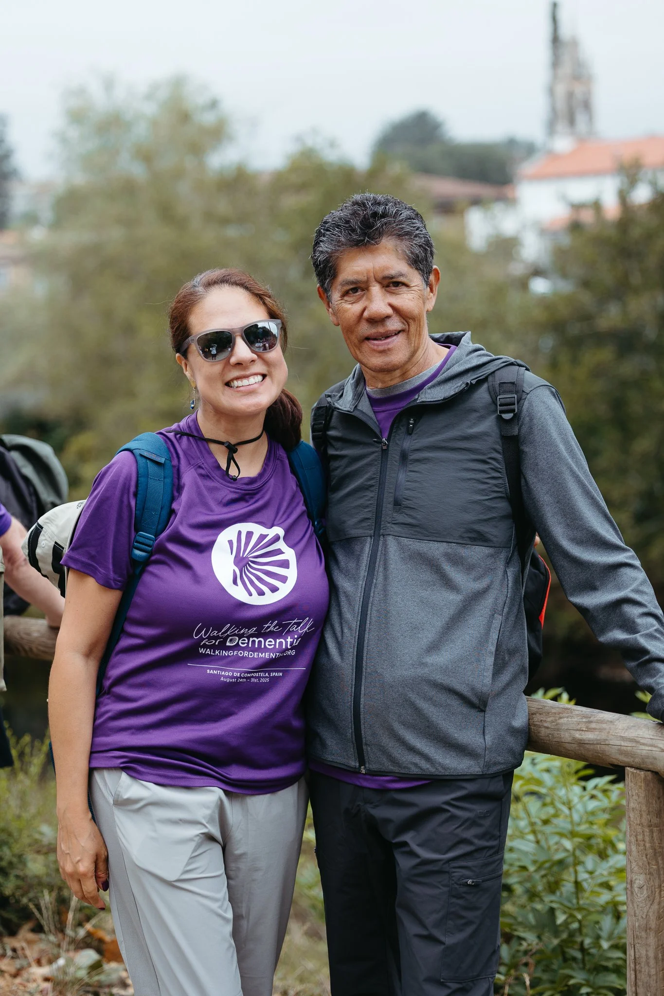 Two people stand outdoors on a trail with trees and houses in the background. The woman on the left is wearing sunglasses, a purple T-shirt with 