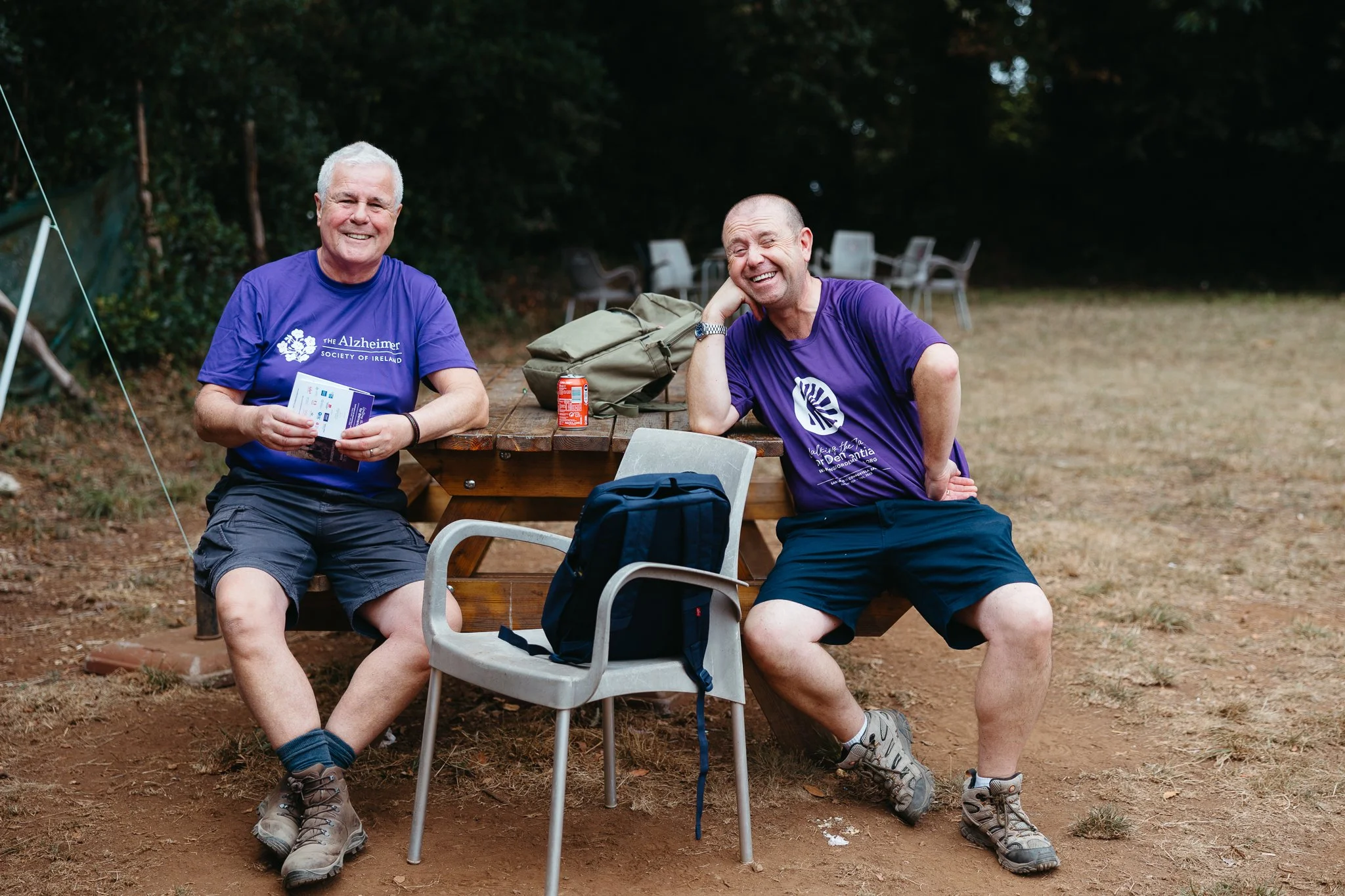 Two smiling men in purple T-shirts, sitting outdoors at a wooden picnic table with a backpack, a can, and a pack of papers. In the background, there are chairs and trees.