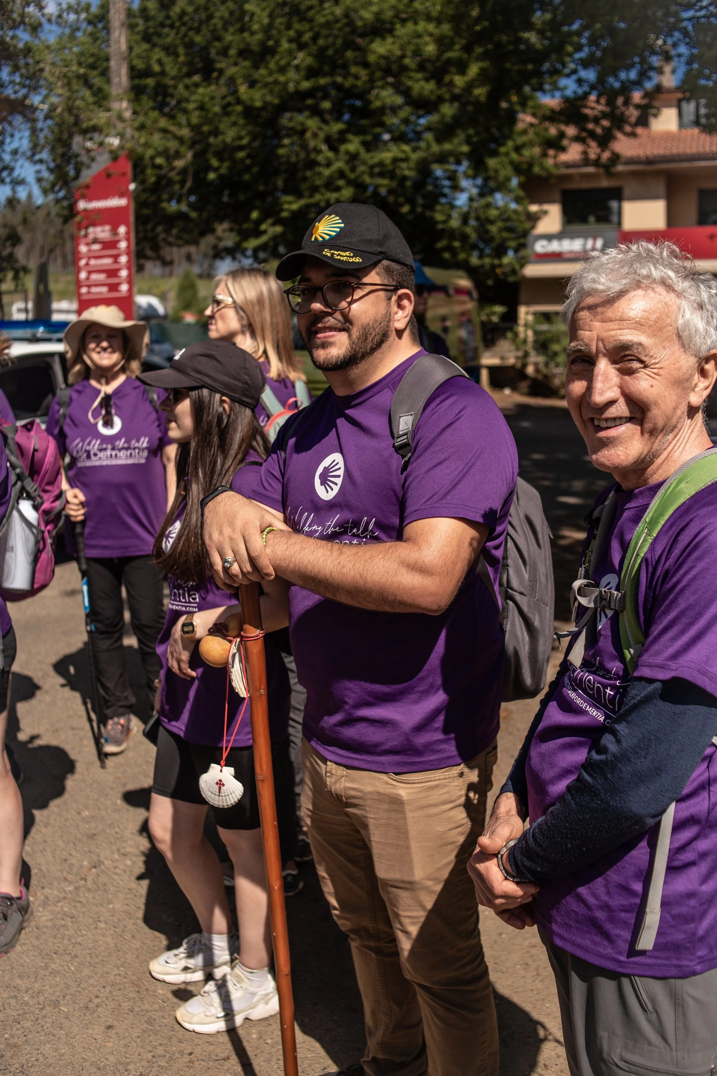 Group of people wearing purple shirts gathered outdoors, some with walking sticks, in sunny weather, with trees and buildings in the background.