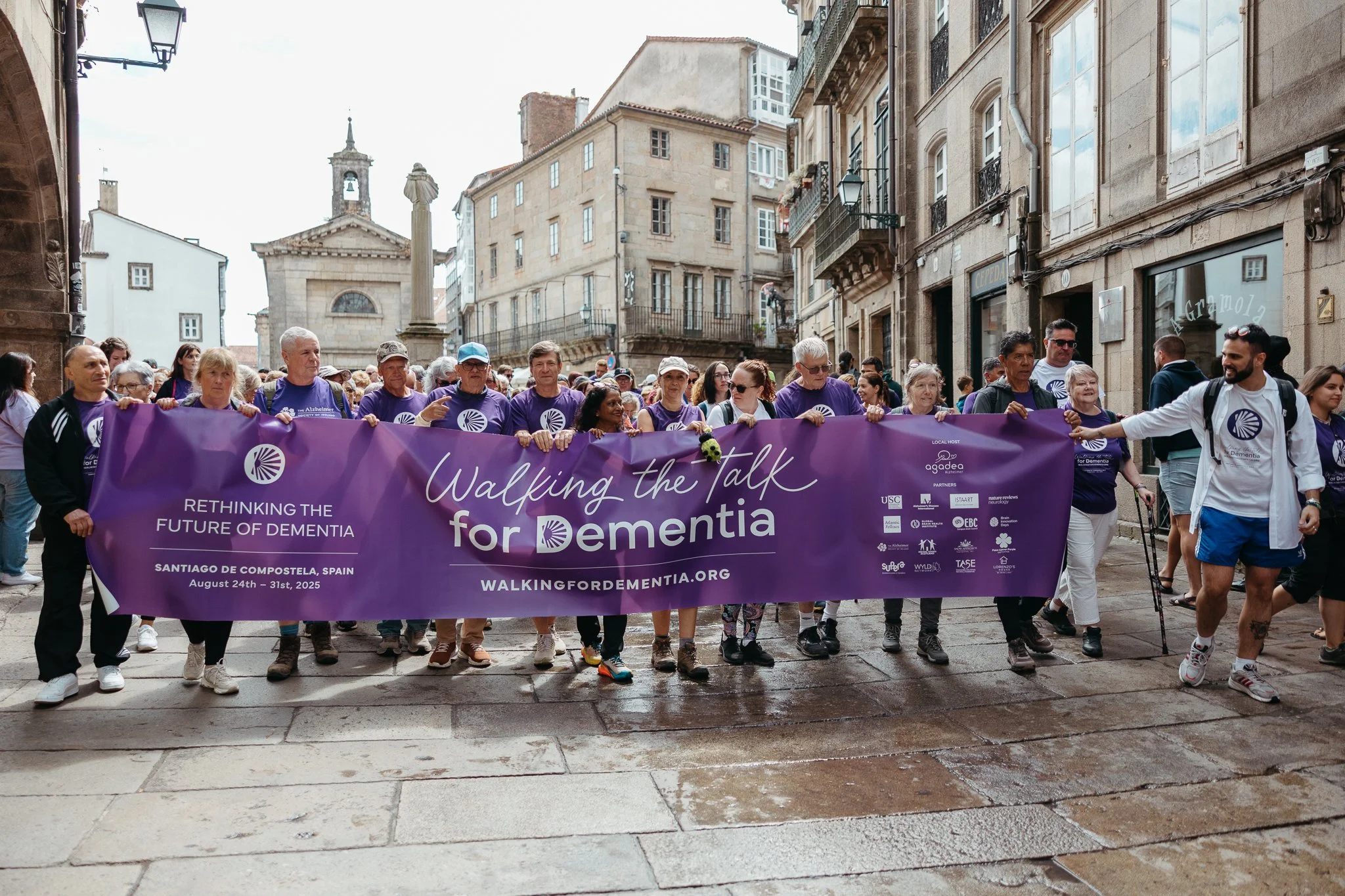 Group of people participating in a dementia awareness march holding a large purple banner that reads "Walking the Talk for Dementia," with the event details in Santiago de Compostela, Spain, from August 24 to 31, 2025.