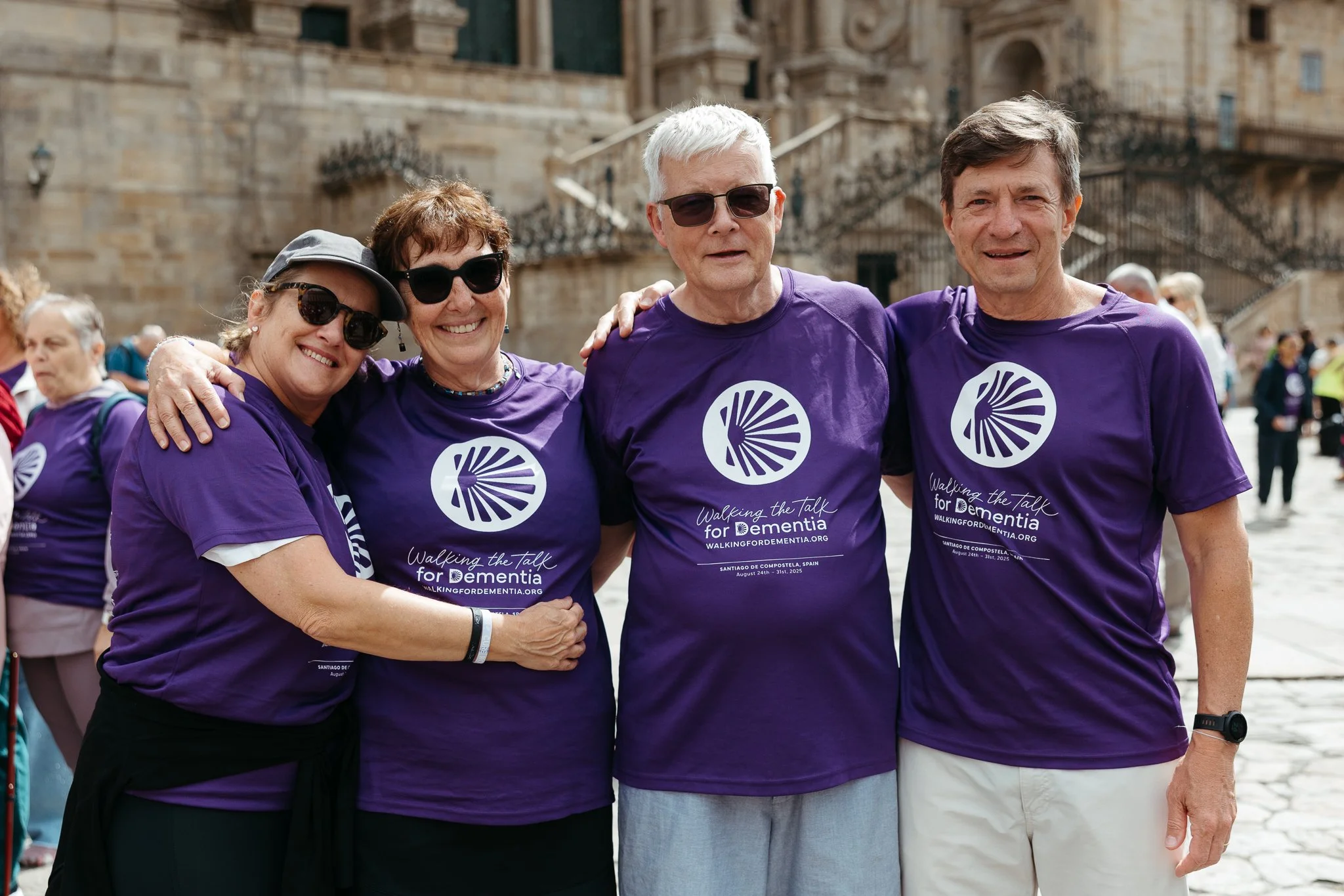 Four adults wearing purple shirts with a dementia awareness logo, standing outdoors, smiling with arms around each other, at a walk event.