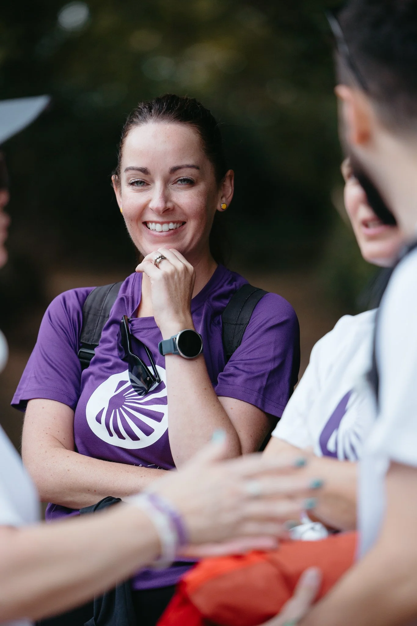 A woman wearing a purple shirt with a logo, smiling and engaging in conversation with others outdoors.