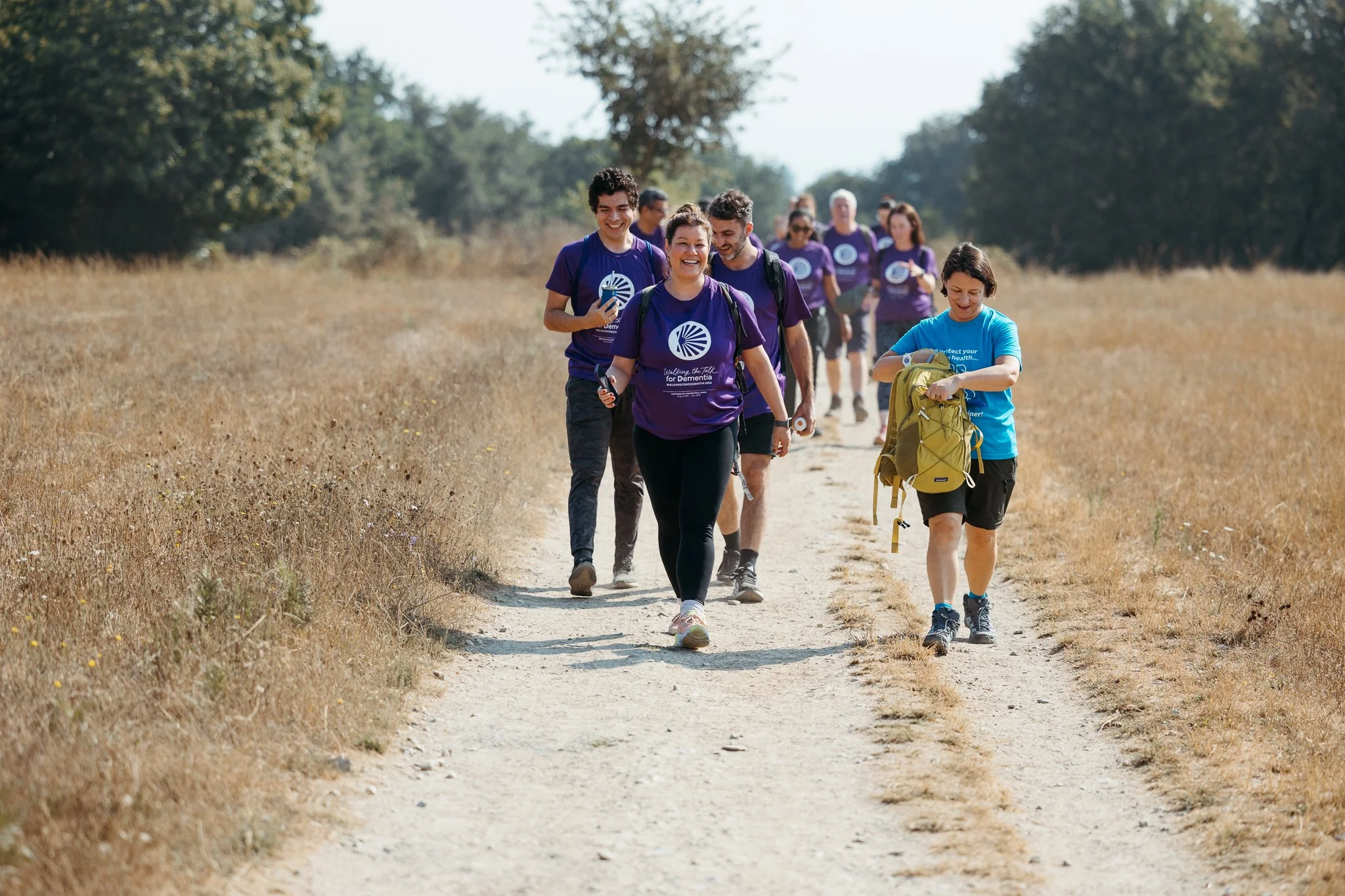 A group of people hiking on a dirt trail through a grassy field with trees in the background, some wearing purple shirts and one girl wearing a blue shirt and yellow backpack.