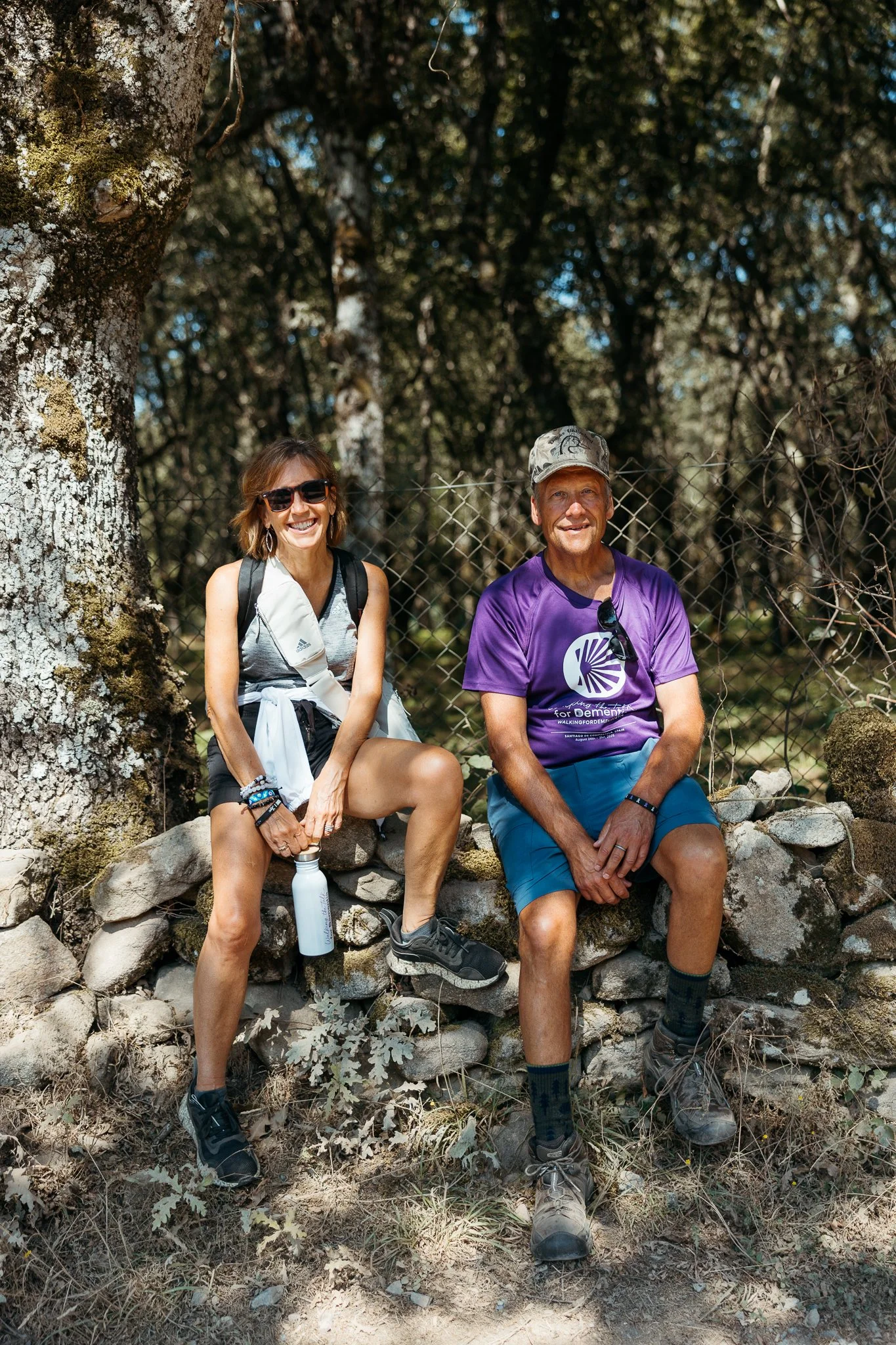 Two people sitting on a stone wall in a wooded area, smiling. The woman on the left is wearing sunglasses, a sleeveless top, and shorts, holding a water bottle. The man on the right is wearing a purple T-shirt, shorts, and a camouflage hat, with sung
