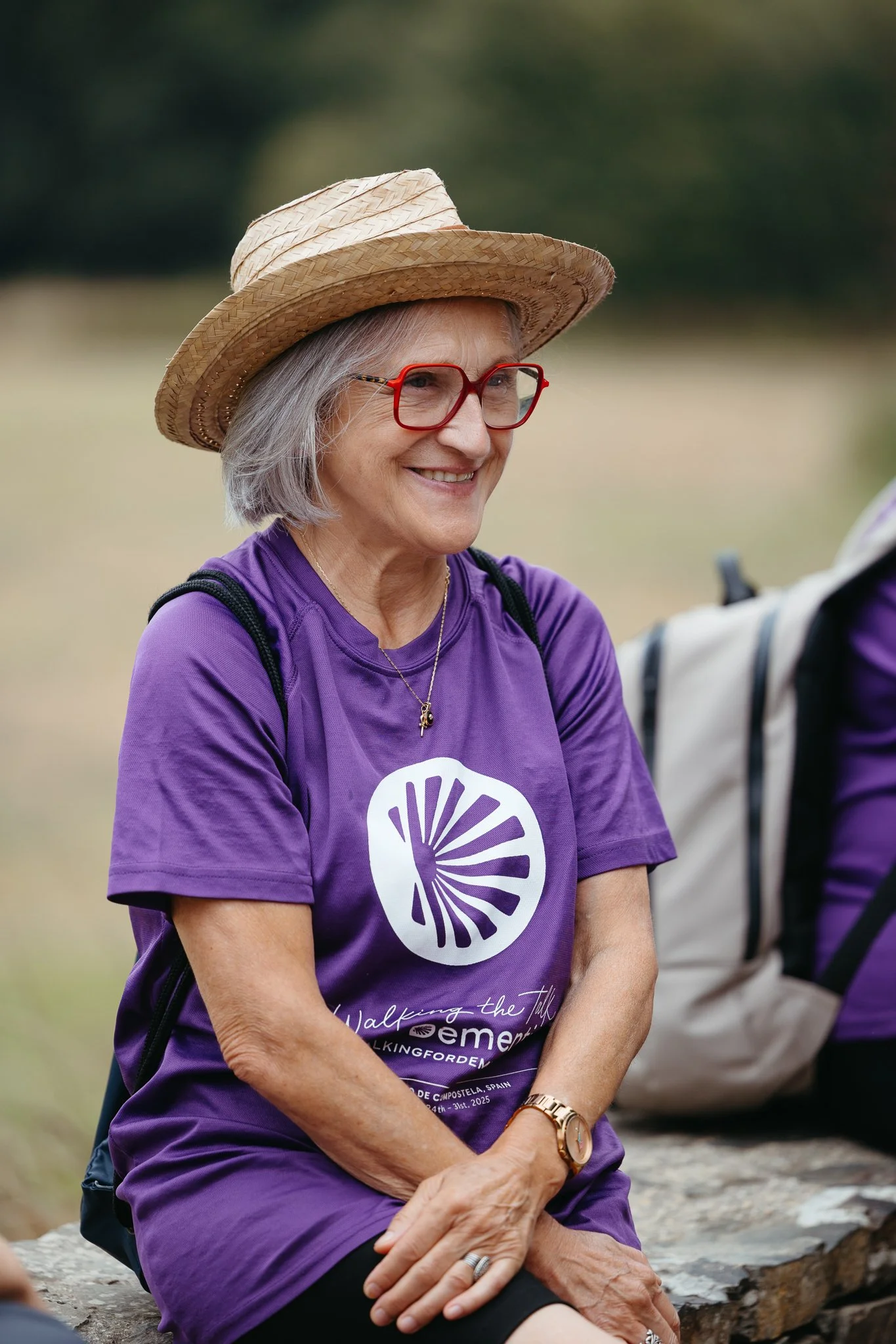 An elderly woman with gray hair, wearing red glasses, a straw hat, a purple T-shirt, a necklace, and a watch, smiling outdoors.