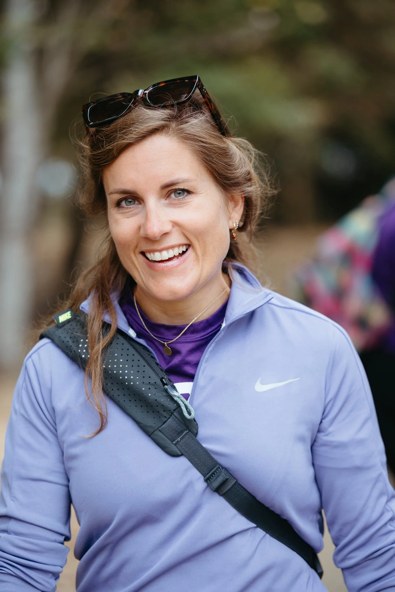 A smiling woman with long brown hair, blue eyes, wearing sunglasses on her head, gold earrings, a purple Nike jacket, and a purple shirt underneath. She has a black crossbody bag and is outdoors with blurred trees in the background.
