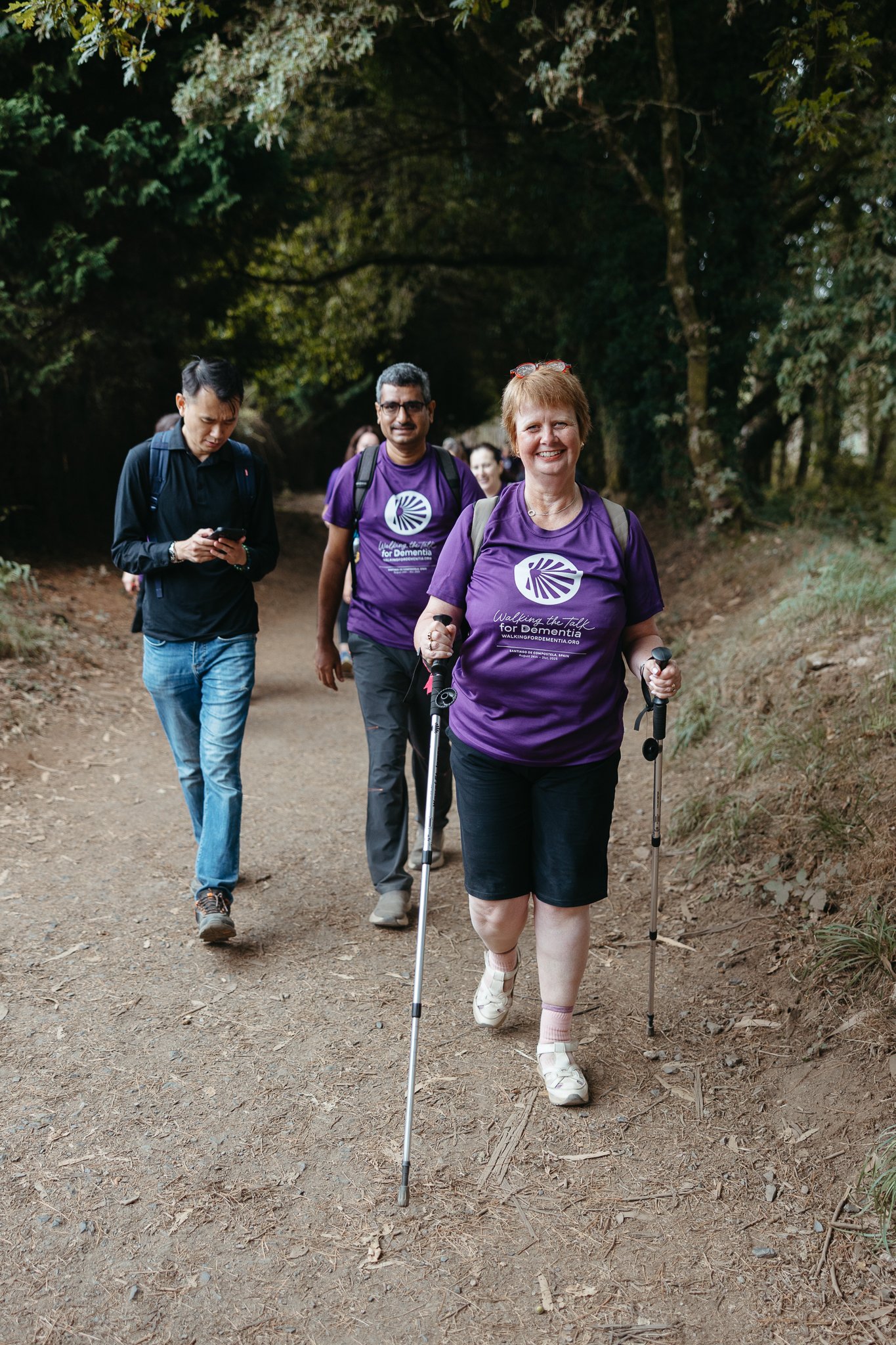 Group of people walking on a dirt trail in a wooded area, including a smiling woman in a purple t-shirt using walking poles, surrounded by other walkers.