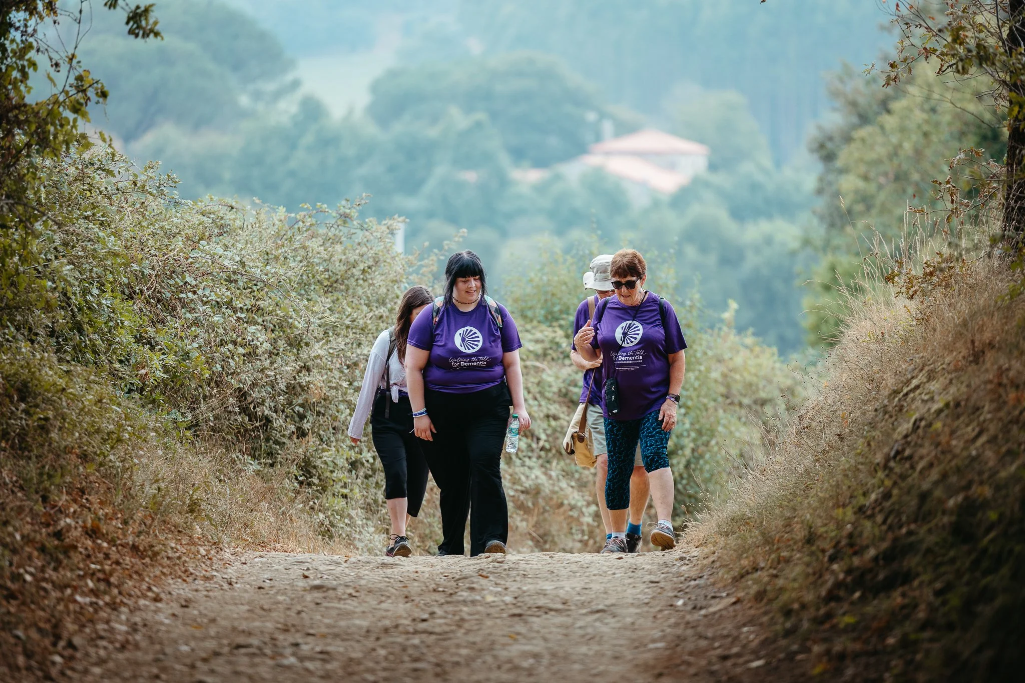 Group of people walking on a dirt trail surrounded by trees and bushes, with a background of hills and buildings.