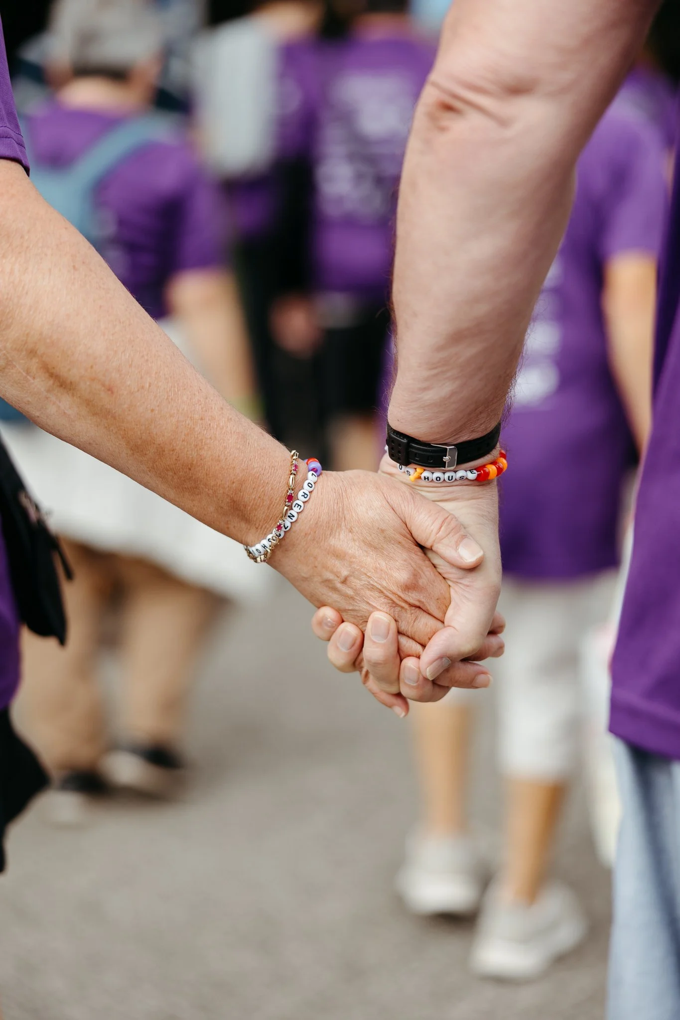 Two people holding hands, with one wearing a bracelet with alphabet beads spelling 'HOPE' and the other with a bracelet spelling 'HOUSES', in a crowd of people wearing purple shirts in the background.