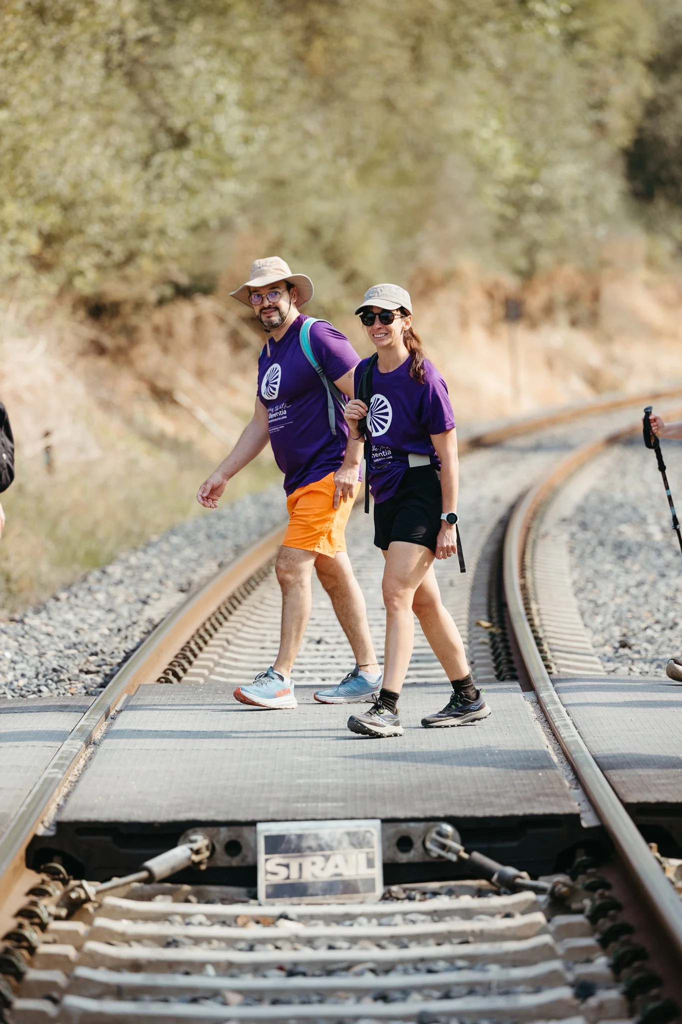 Two people walking on a railroad track outdoors, wearing purple T-shirts, hats, and sunglasses, smiling and enjoying a hike, with a third person partially visible on the right holding a hiking pole.