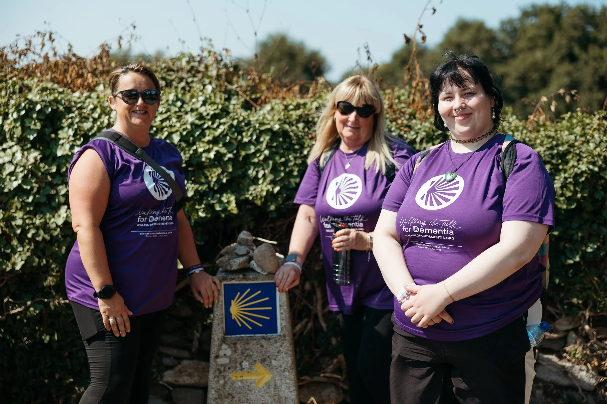 Three women wearing purple T-shirts with a logo and text, standing outdoors next to a stone marker with yellow shell symbol and arrow, smiling at the camera during a walk for dementia awareness.