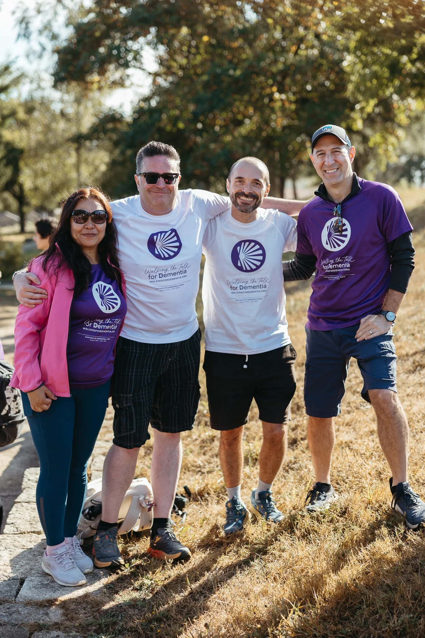 Group of four people standing outdoors on a sunny day, participating in a walk for dementia awareness. They are smiling, wearing casual athletic clothing, with three wearing matching t-shirts with a shell logo and the text 'Walking the Talk for Demen