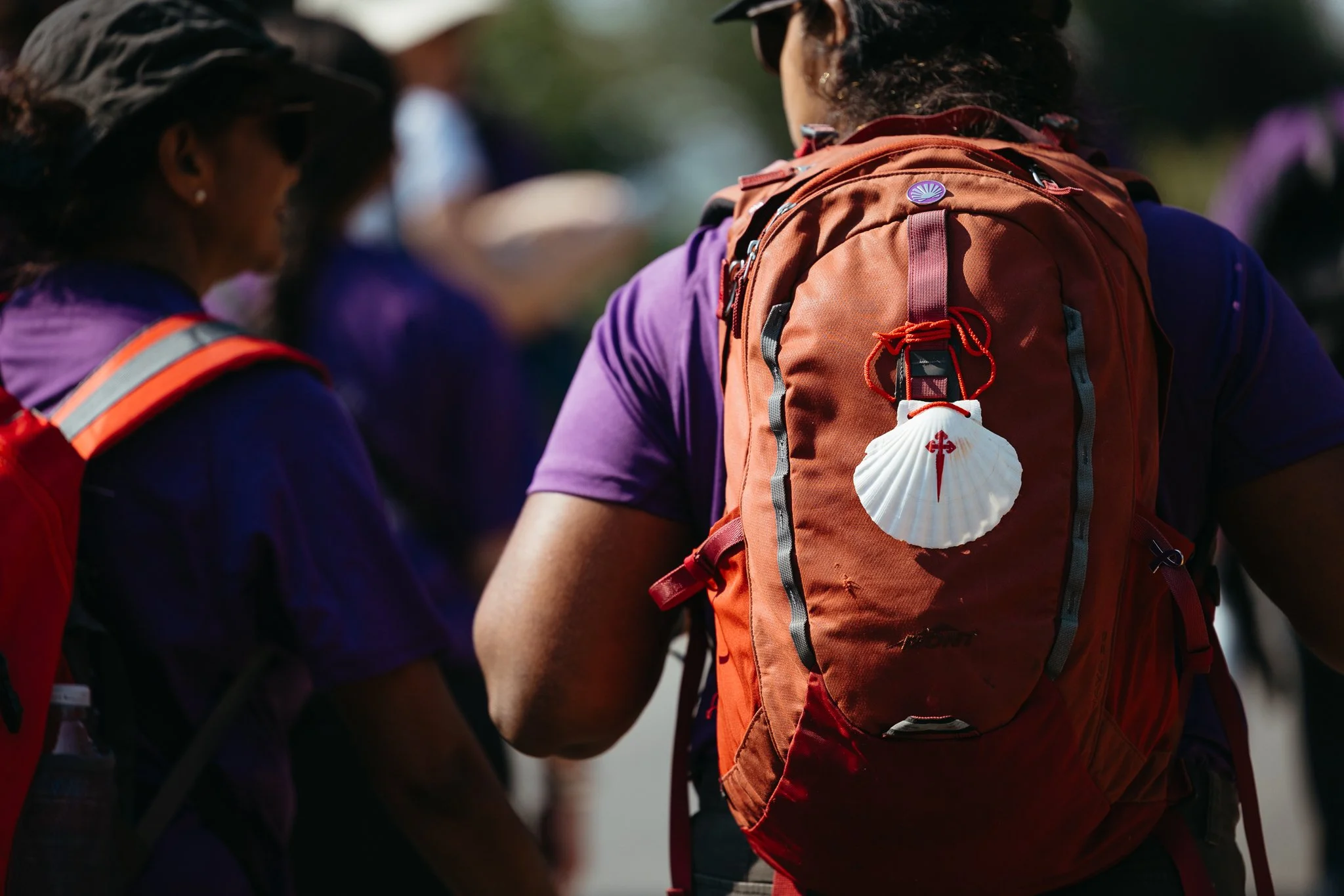 A group of hikers gathered outdoors, with a focus on a person wearing an orange backpack with a white scallop shell and a small red cross attached to it.