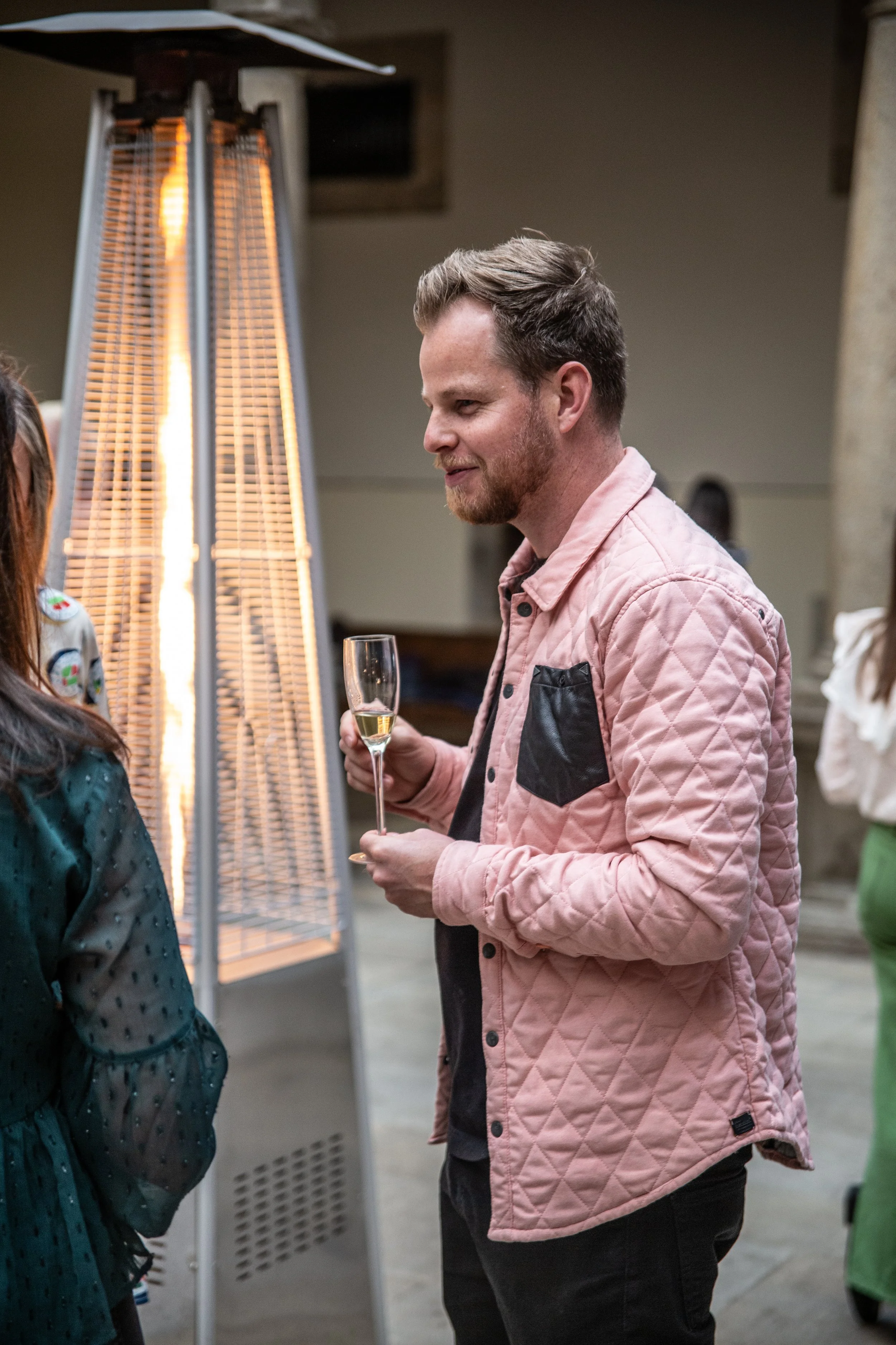 A man holding a glass of champagne at a social gathering, standing near a tall outdoor heater.