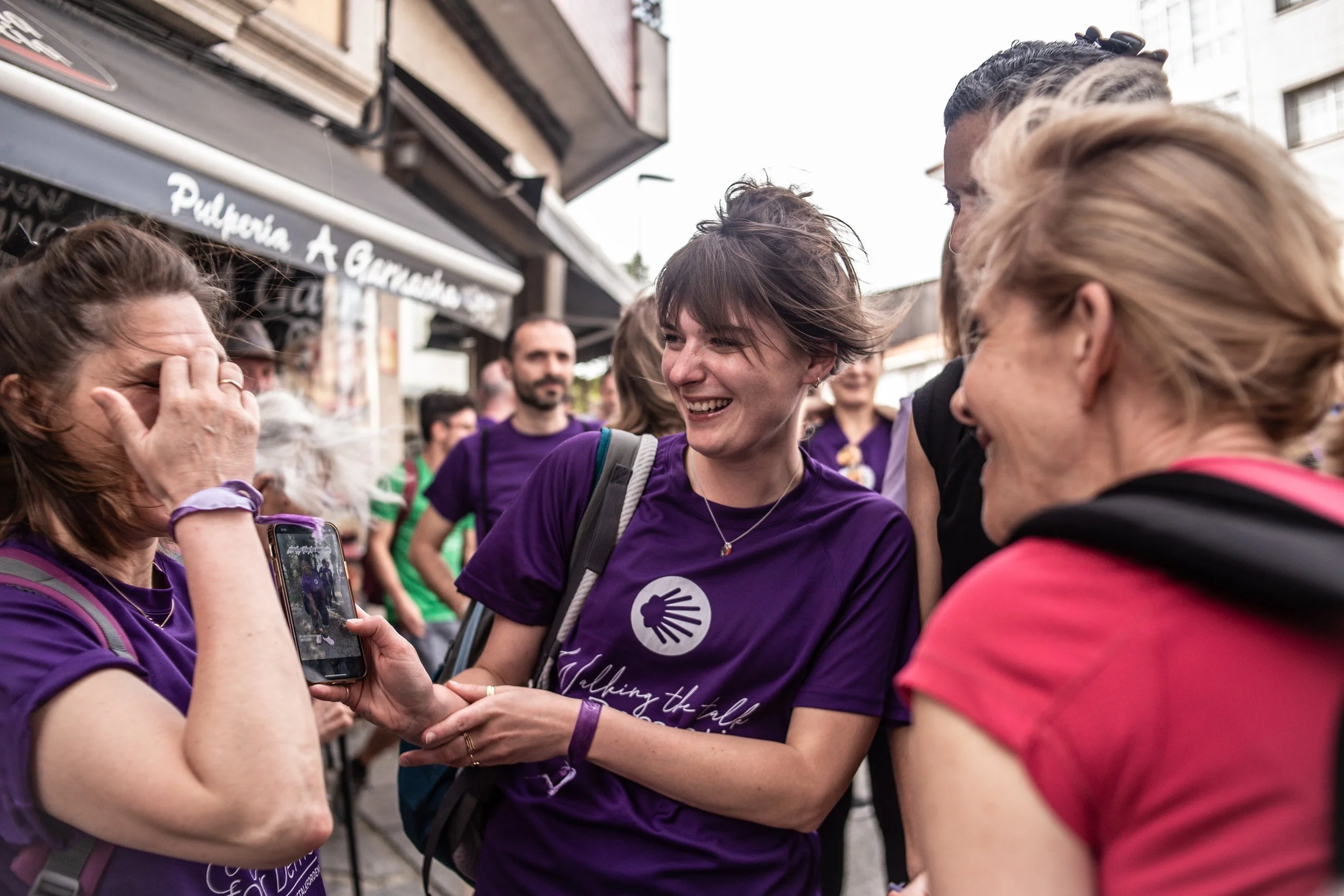 Group of people, mostly women, socializing outdoors in a busy street scene, some wearing purple T-shirts, smiling and engaging with each other.
