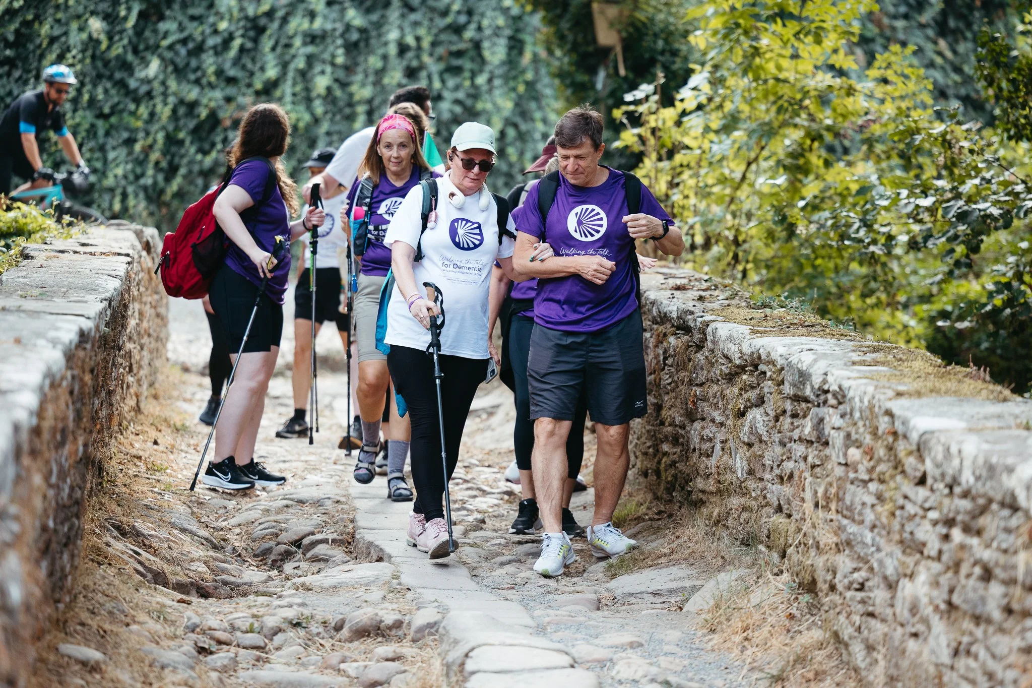 Group of people hiking on a trail with stone walls, surrounded by trees, some wearing purple shirts and using trekking poles.