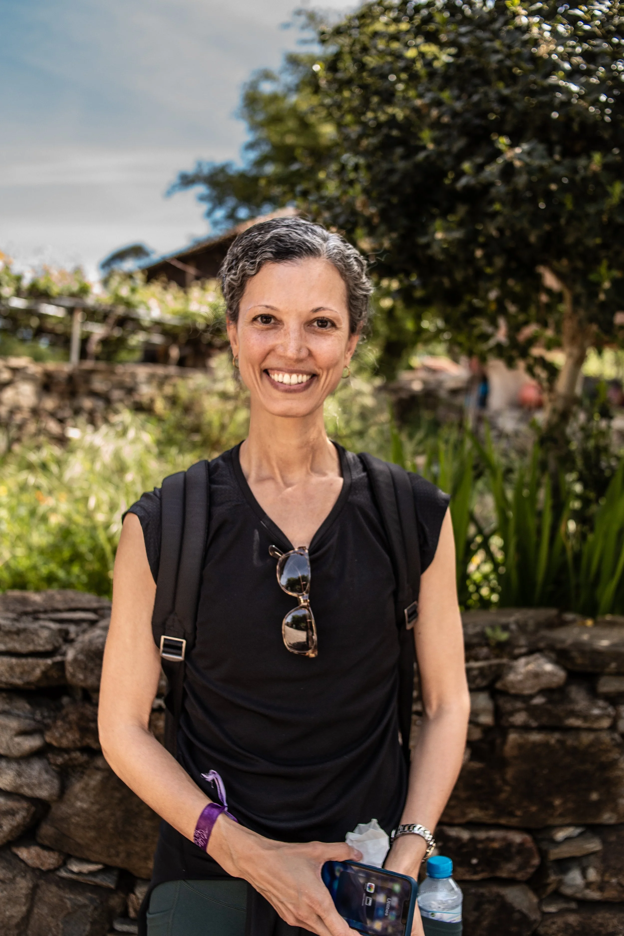 Smiling woman with short gray hair wearing a black sleeveless top, sunglasses hanging from her shirt, a wristband, and a watch, holding a water bottle and a phone, outdoors with trees and a stone wall in the background.