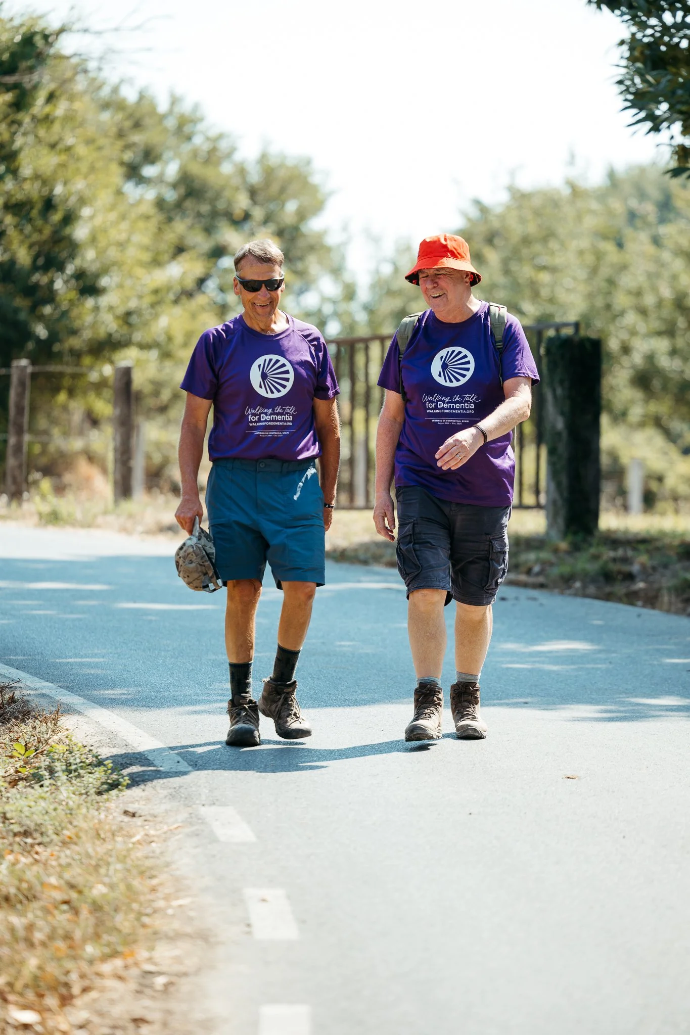 Two middle-aged men walking and talking along a paved path in a park, both wearing purple t-shirts supporting a dementia awareness walk, with trees and a metal fence in the background.