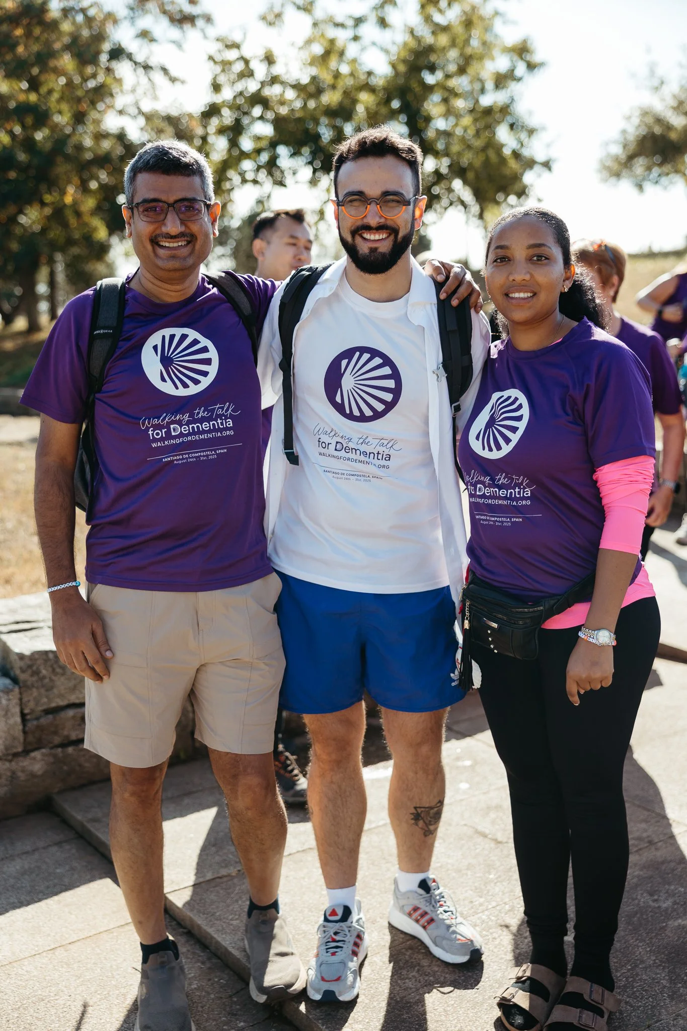 Three smiling people standing outdoors, wearing matching purple t-shirts with a logo and the text 'Walking the Talk for Dementia,' participating in a walk event supporting dementia awareness, with trees and other participants in the background.