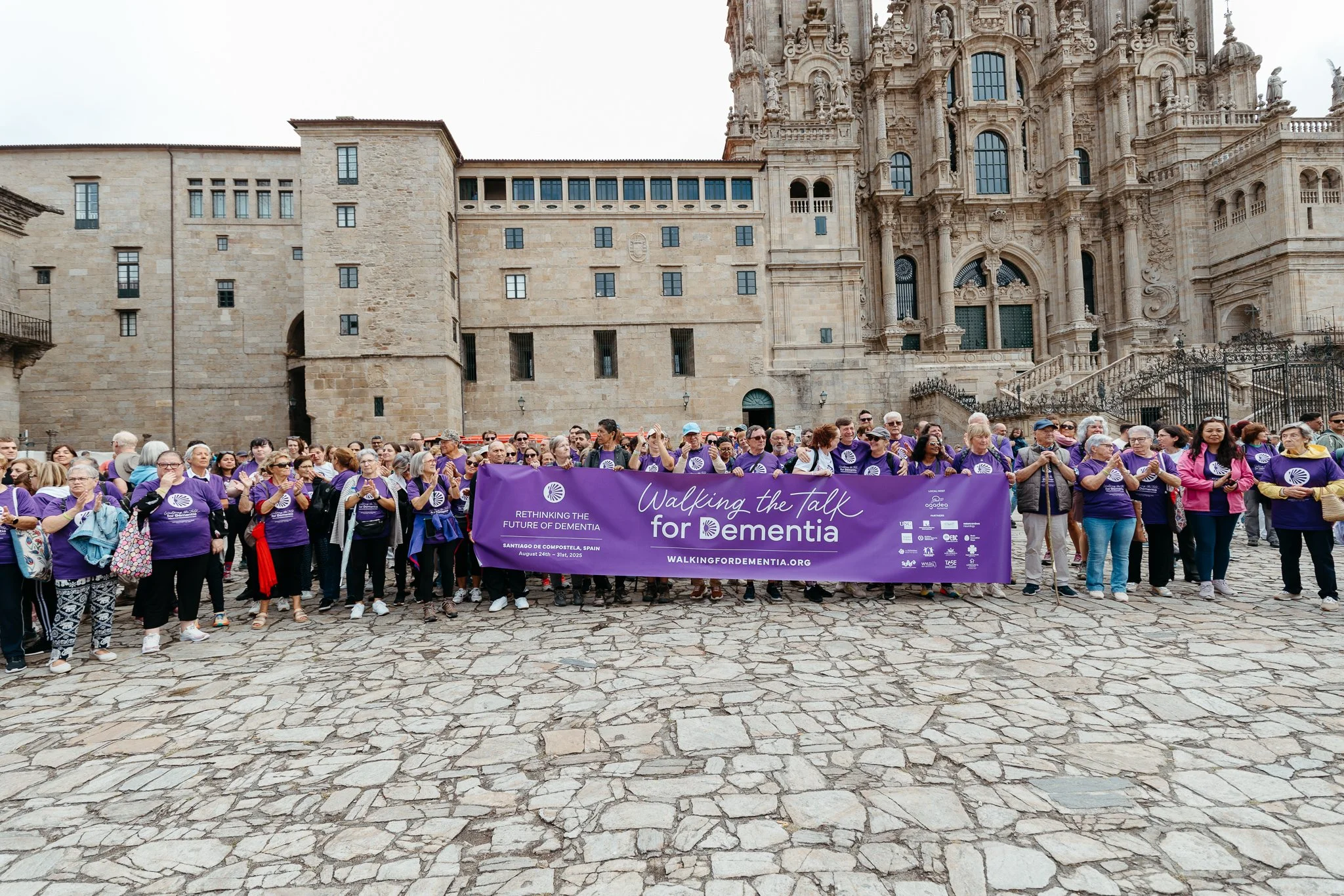 Group of people participating in a dementia awareness march, holding a large purple banner that reads "Walking the Talk for Dementia." They are gathered in front of a historic building with ornate architecture and stone steps.