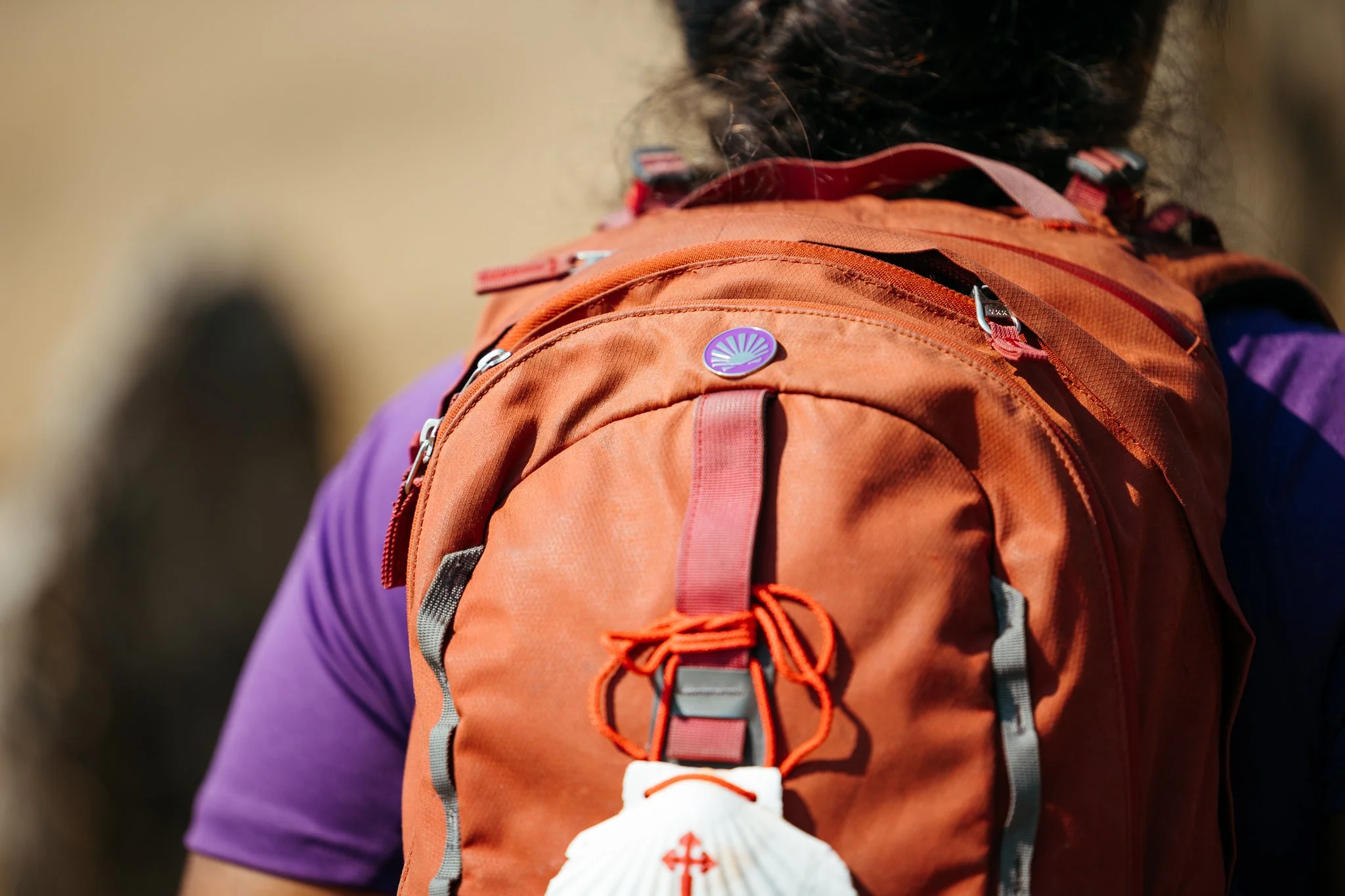 Close-up of a person wearing an orange hiking backpack with a purple pin, a white seashell attached to the front, and purple shirt visible on the shoulder.