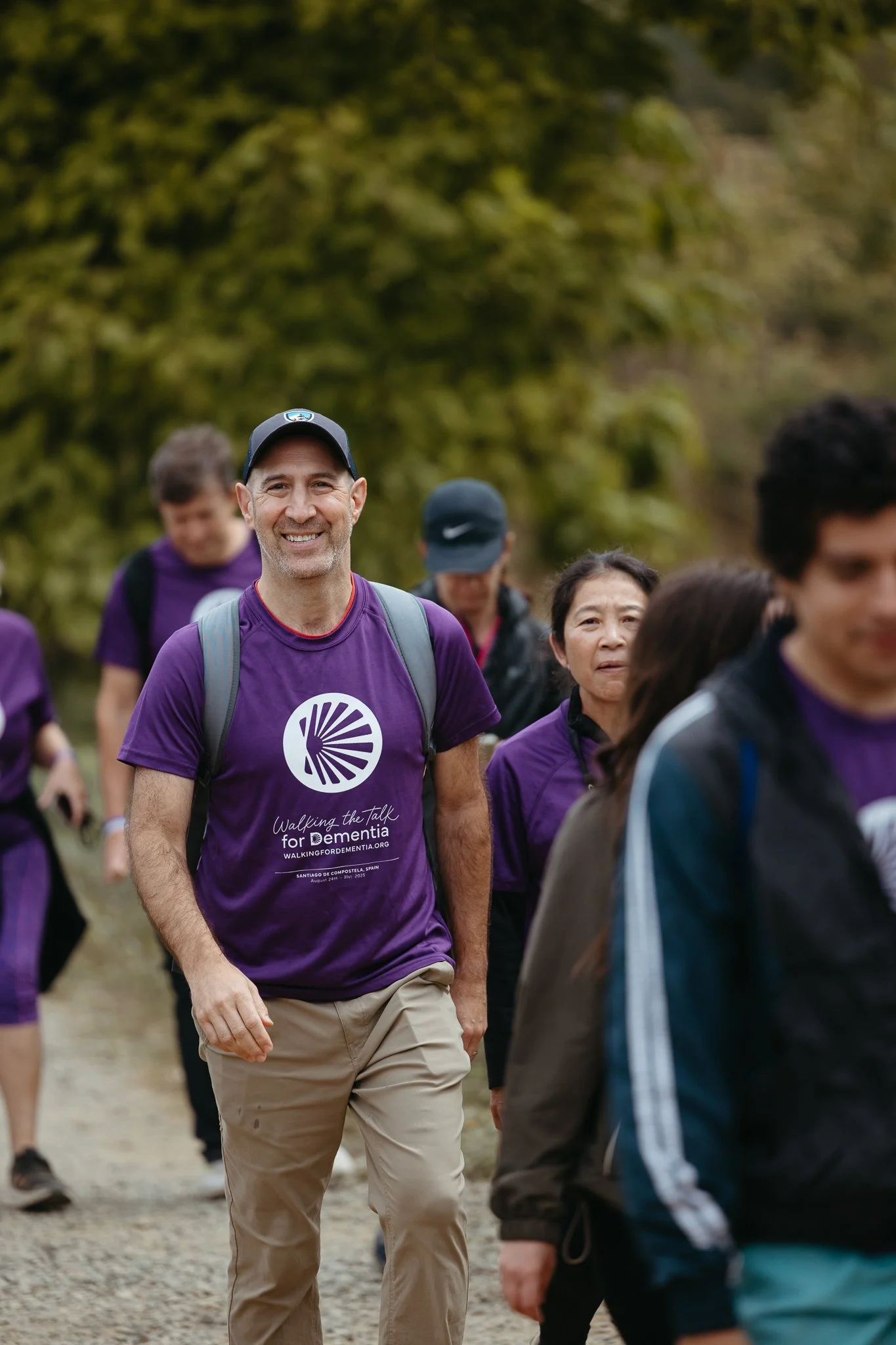 People walking outdoors, wearing purple shirts for an awareness event, with one smiling man in focus.