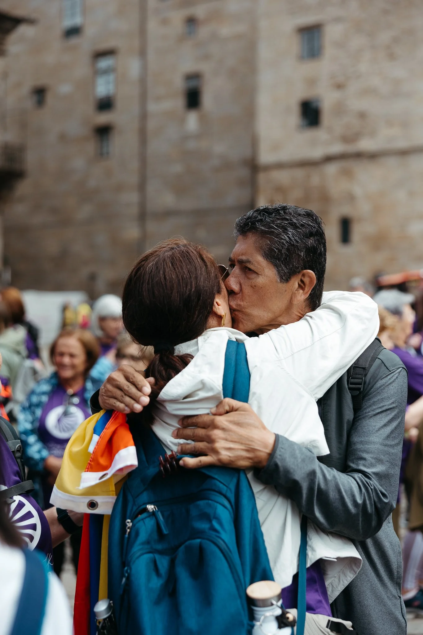 A man and woman embrace and kiss in a crowd, with the woman wearing a rainbow flag draped over her shoulder.